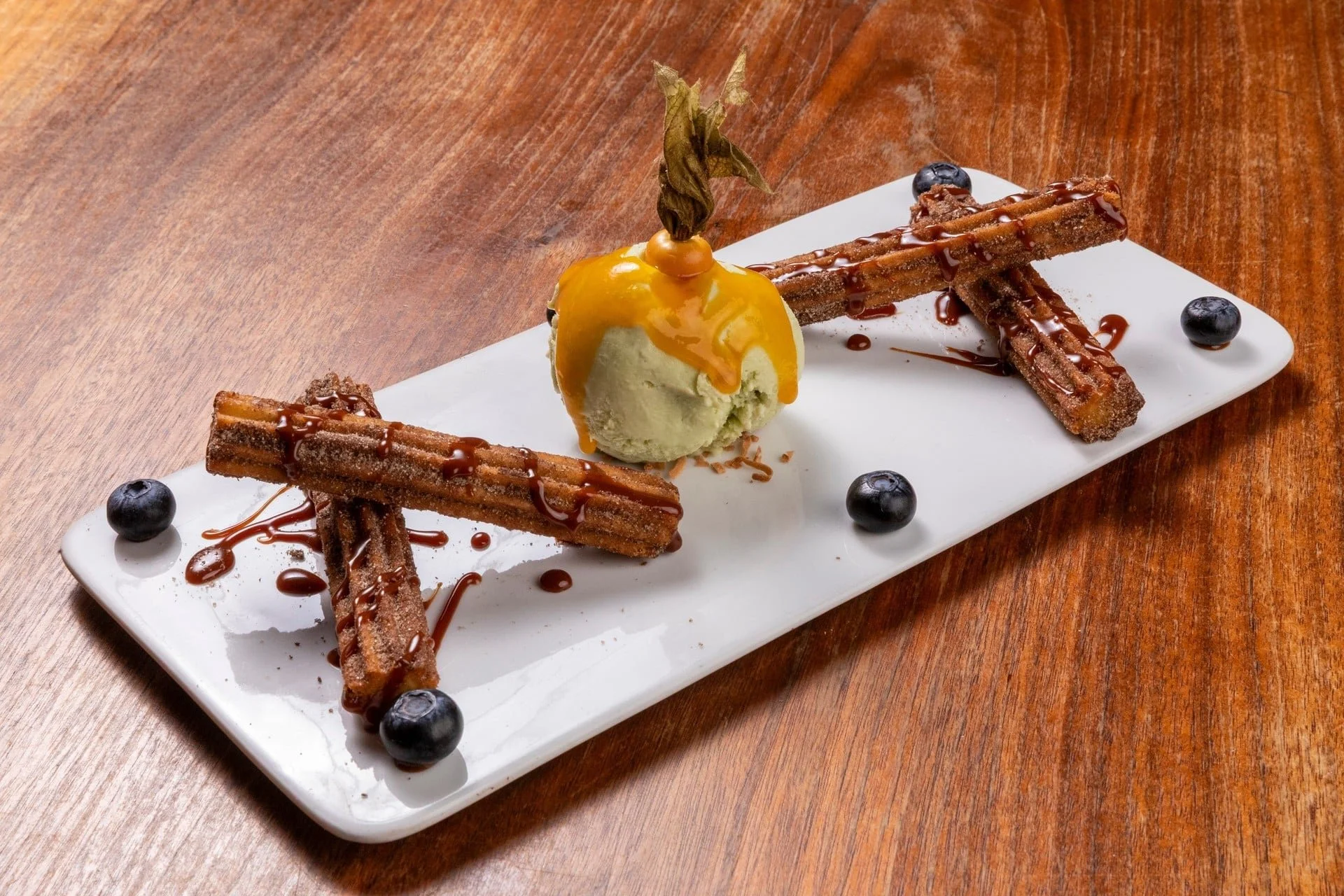 Plate of churros drizzled with chocolate sauce, served with a scoop of vanilla ice cream topped with caramel sauce and garnished with a leaf, surrounded by blueberries on a white rectangular plate on a wooden table.