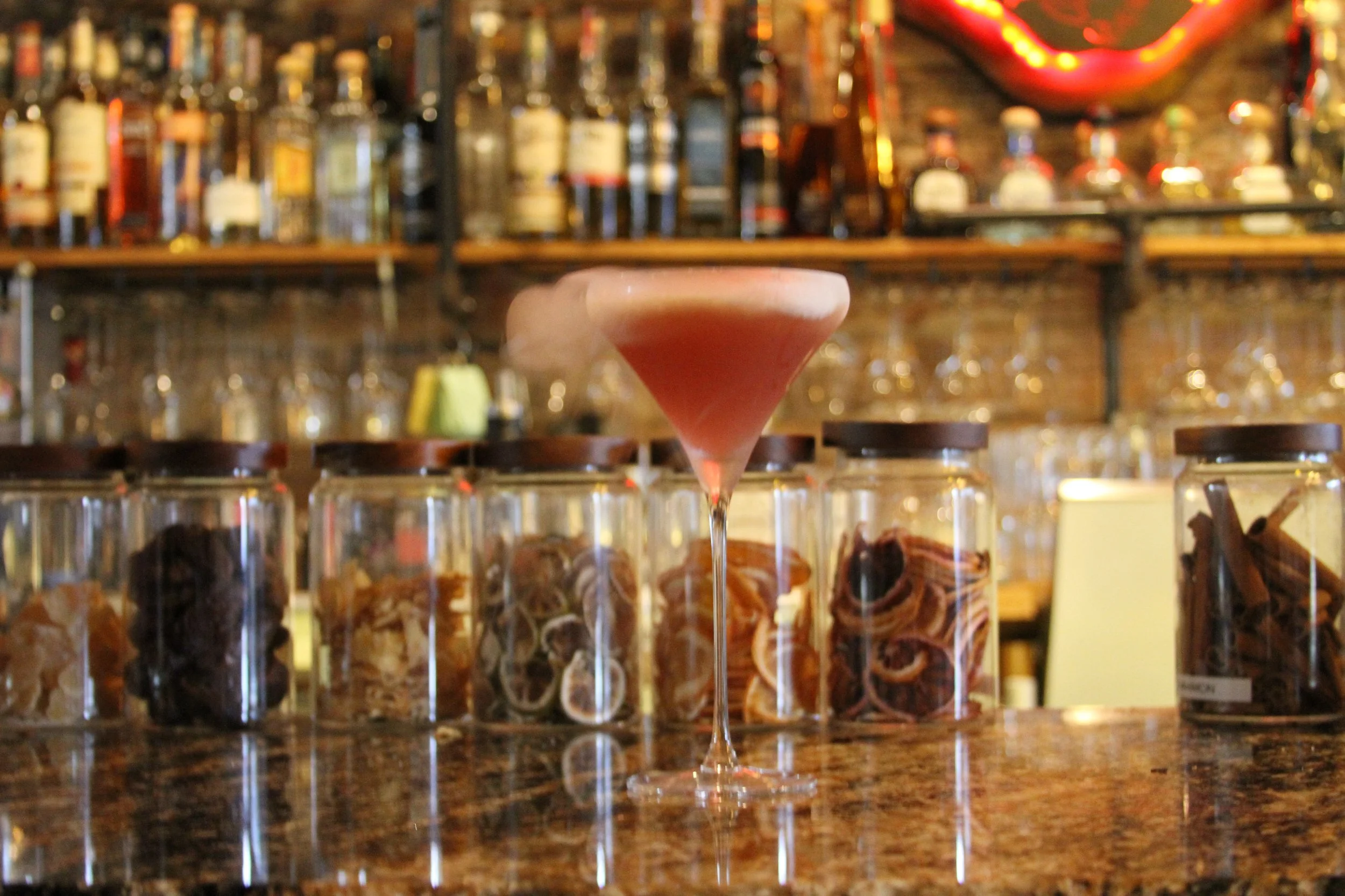 A pink cocktail in a martini glass is placed on a bar counter with glass jars filled with snacks or candies underneath. The background shows shelves with various bottles of alcohol and ceiling lights, creating a cozy bar atmosphere.
