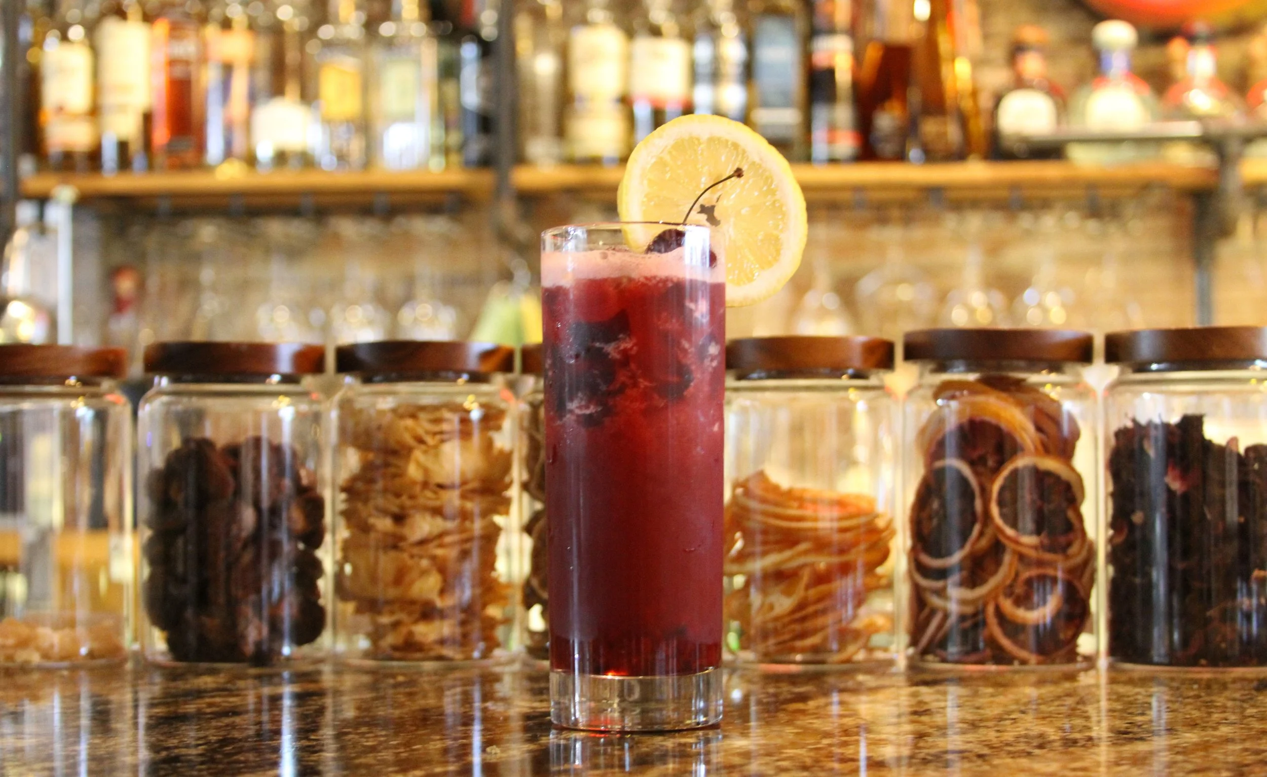 A tall glass of red fruit juice garnished with a lemon wheel and a cherry, set on a bar counter with jars of cookies or snacks behind it.