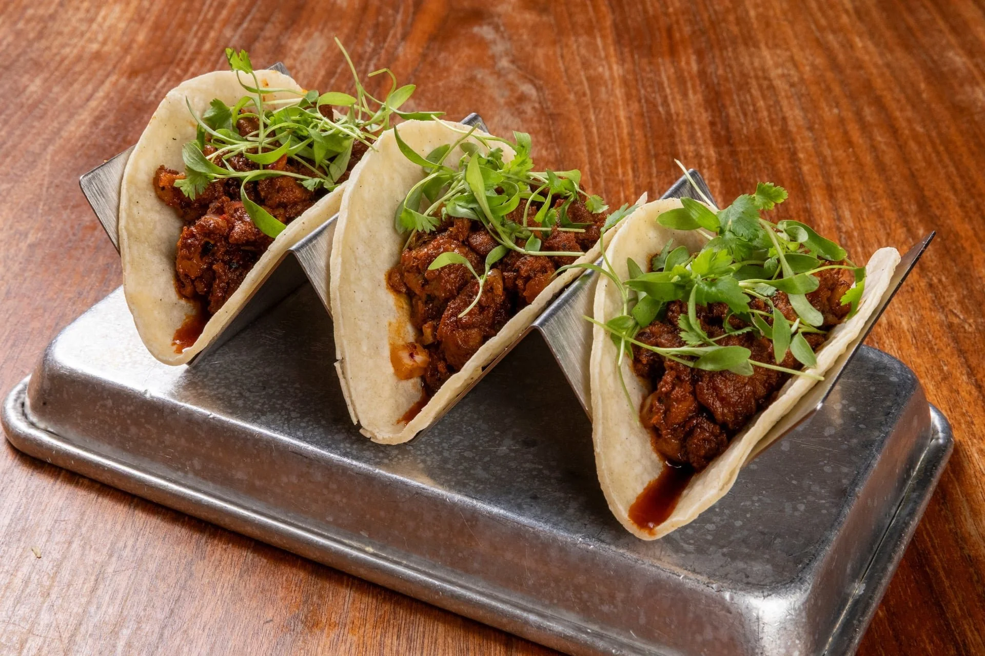 Three tacos filled with seasoned meat, topped with microgreens and cilantro, served on a metal taco holder.