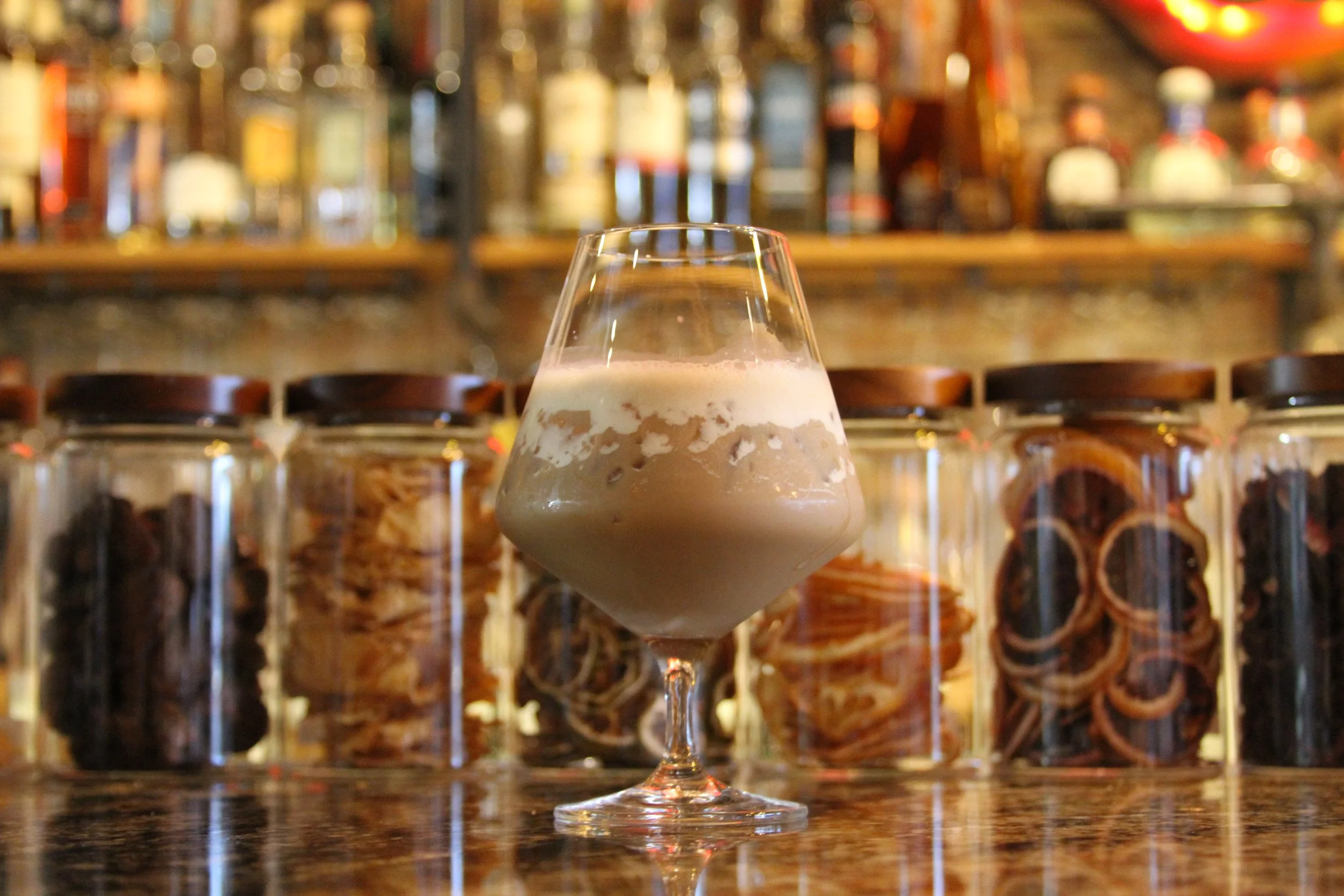 A glass of creamy dessert on a wooden surface in front of a blurred background of liquor bottles on shelves.