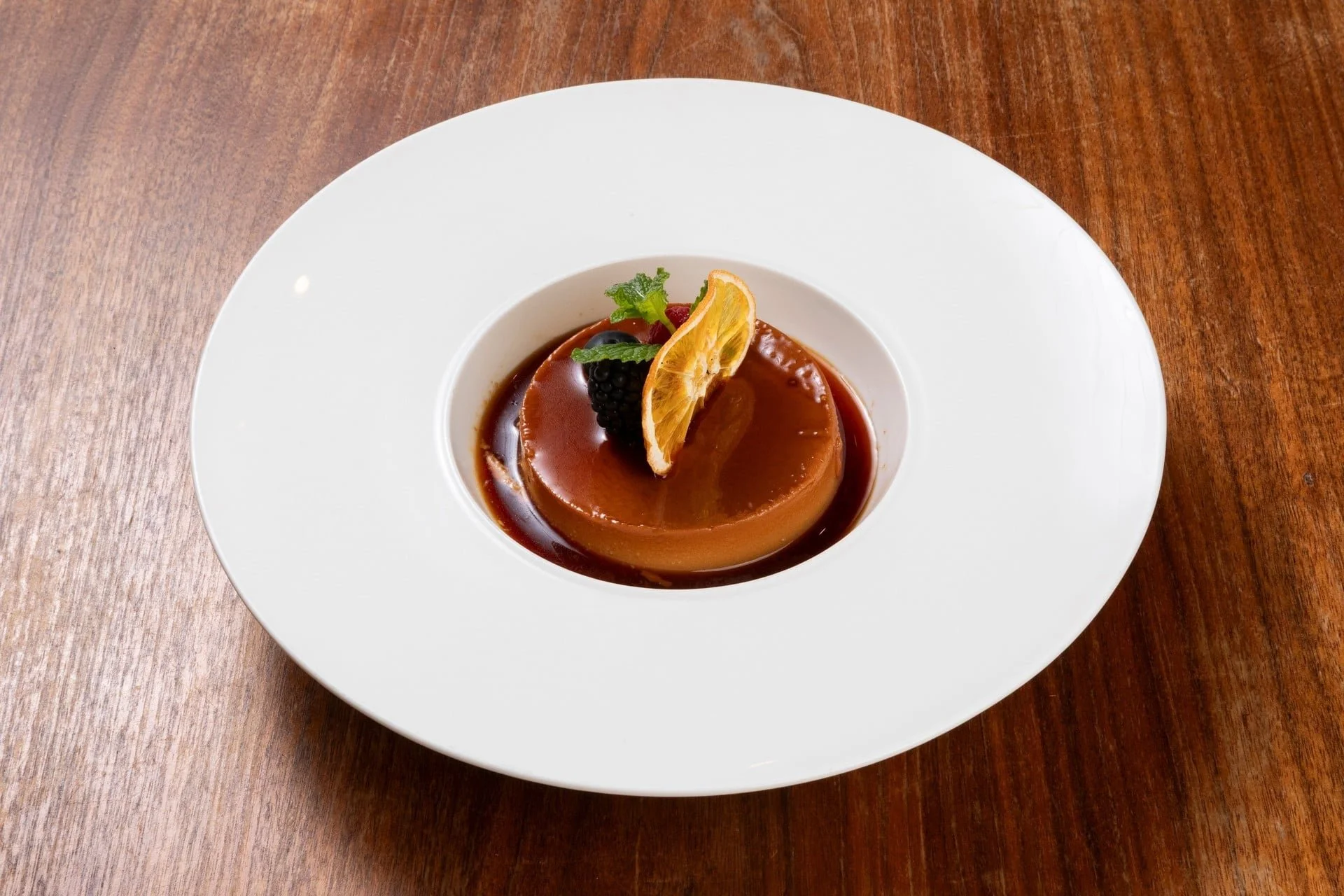 Caramel flan dessert garnished with a lemon slice, fresh mint, and berries on a white plate, placed on a wooden table.