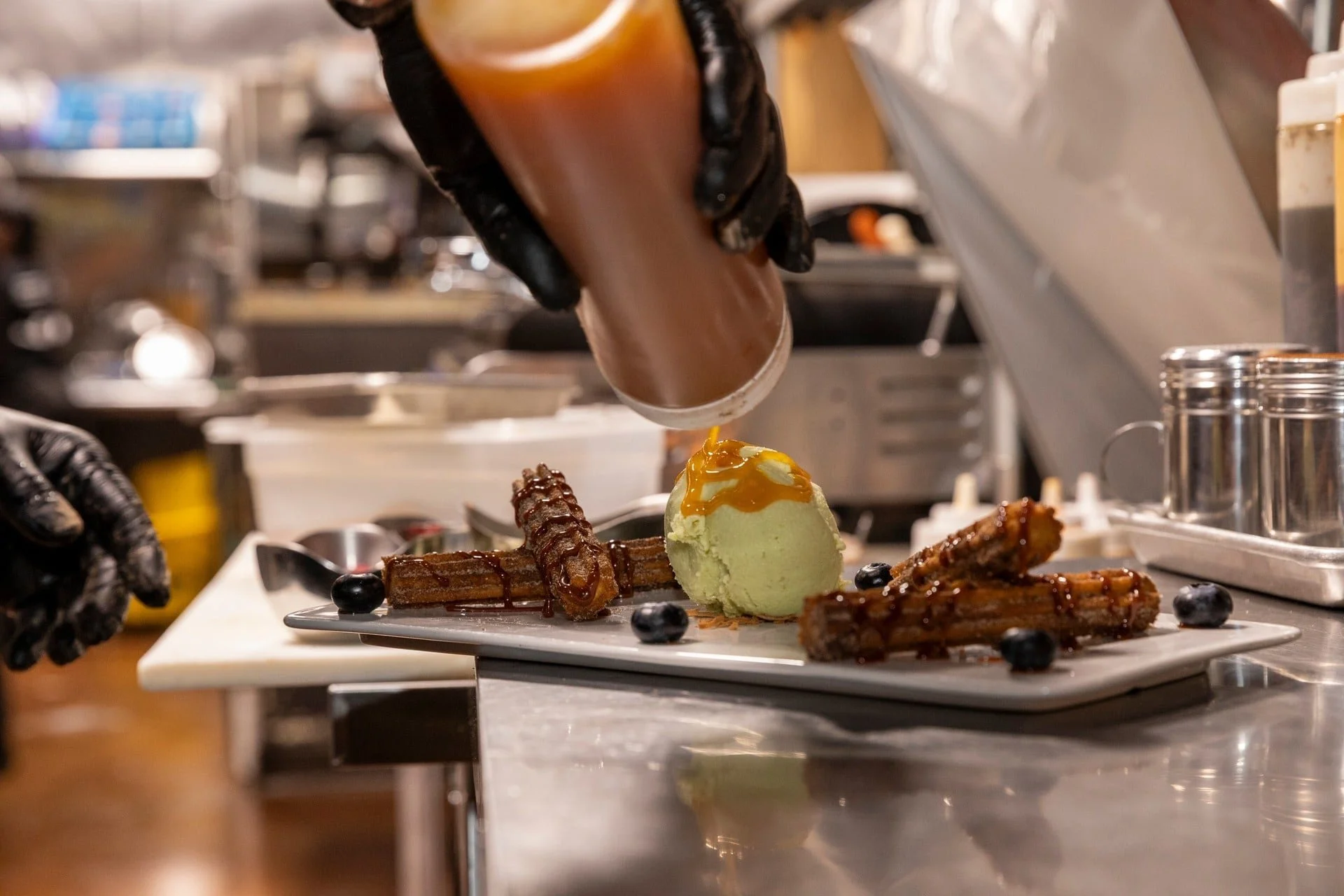 A chef zips caramel syrup over a plate with waffles, a scoop of green ice cream, and blueberries in a professional kitchen.