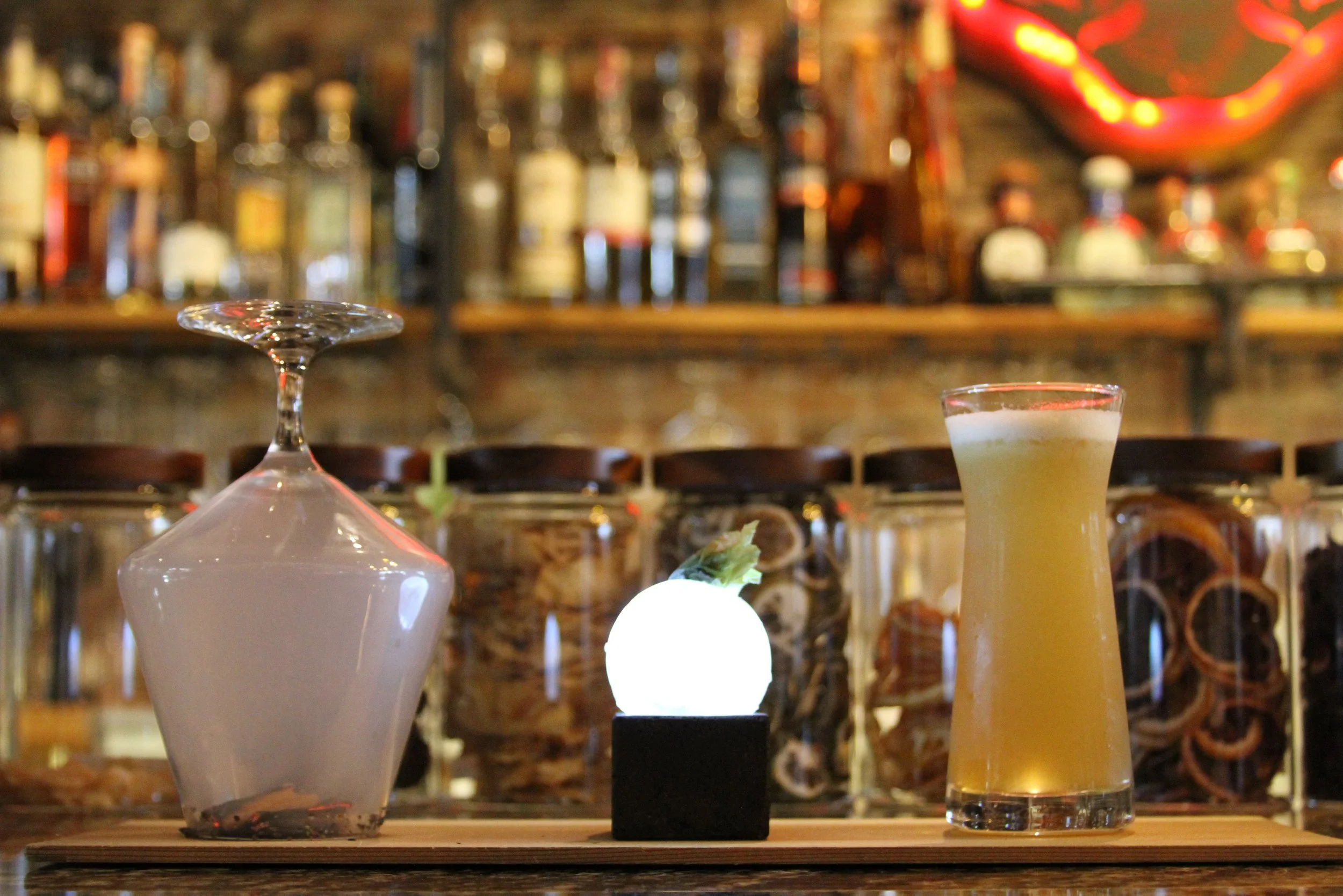 A blurred bar scene with bottles on shelves in the background, and a clear martini glass, yellow cocktail, and illuminated decoration on the bar counter in the foreground.