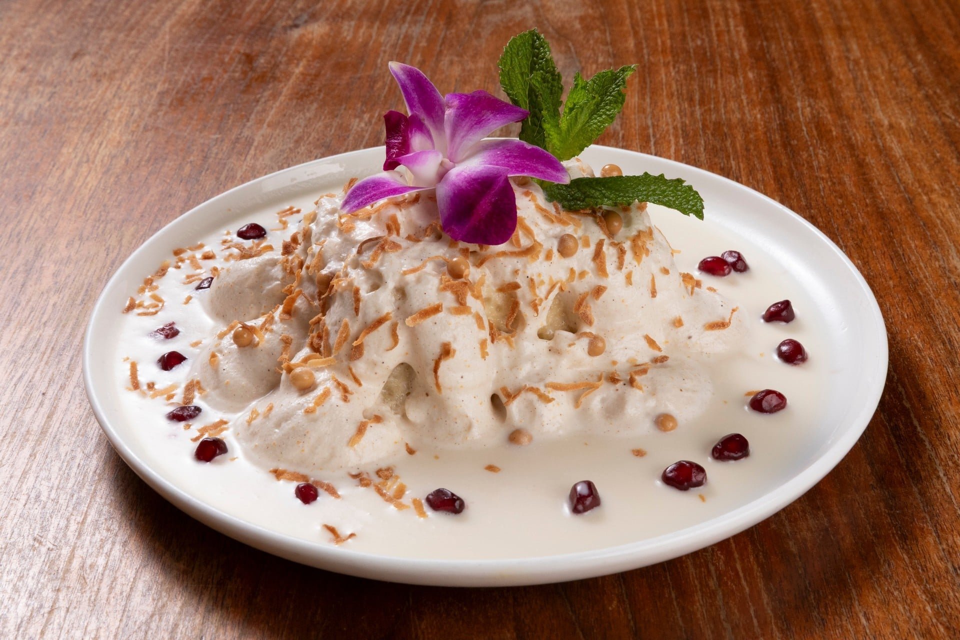 A plate of Thai dessert, coconut ice cream garnished with edible orchid, mint leaves, toasted coconut flakes, and pomegranate seeds on a wooden table.