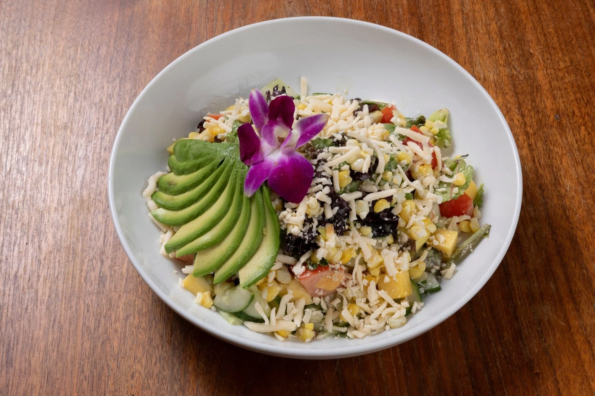 Bowl of mixed salad with sliced avocado, shredded cheese, cherry tomatoes, corn, black beans, and an edible flower on top, on a wooden table.