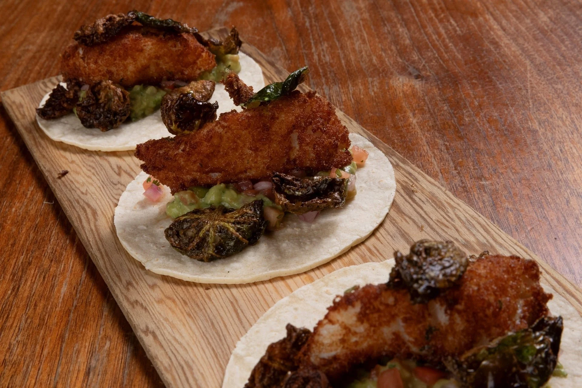 Close-up of fish tacos topped with fried fish, grilled jalapeños, and vegetables on small tortillas on a wooden serving board.
