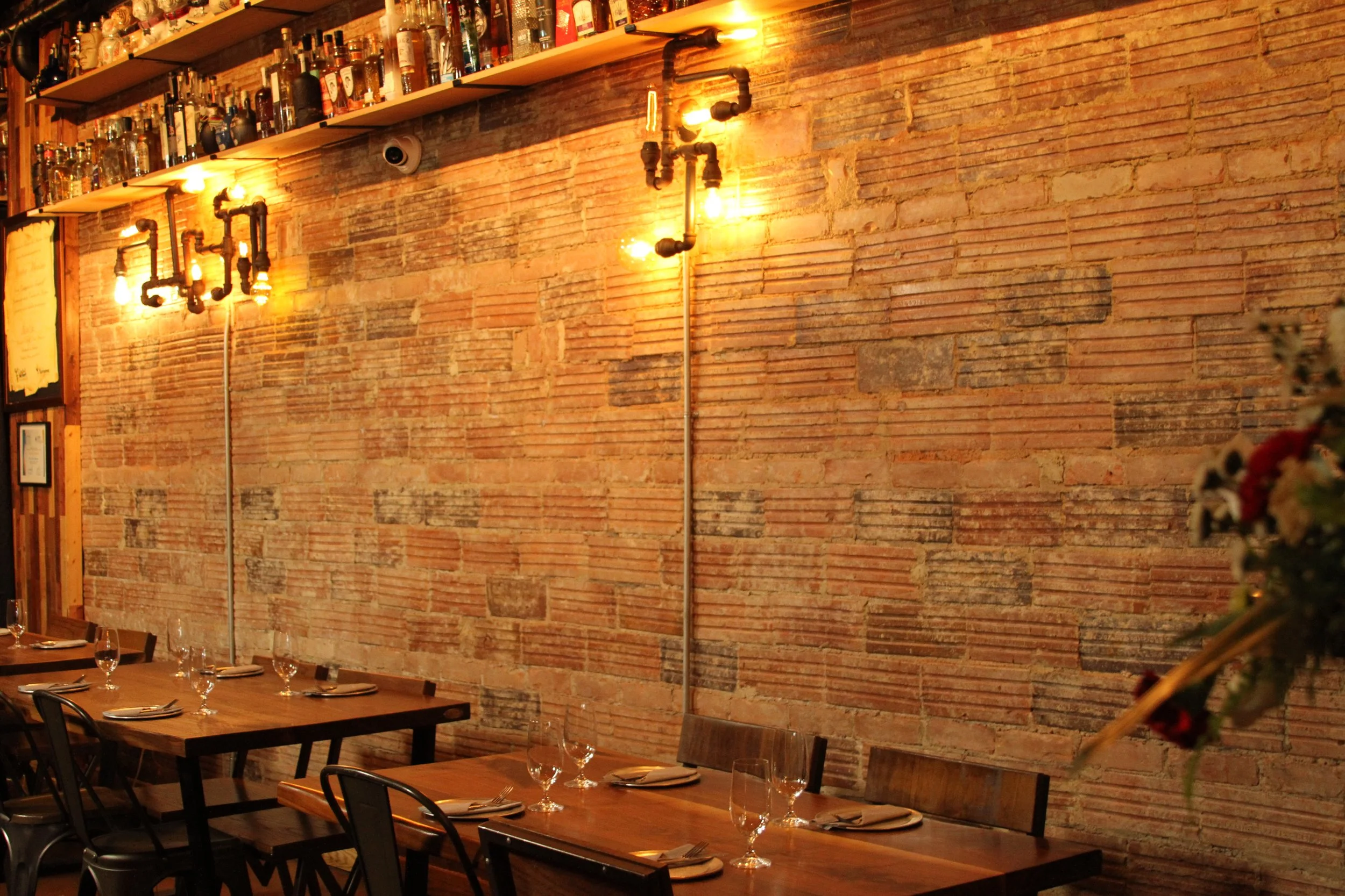 A cozy restaurant with a rustic brick wall, decorated with shelves of liquor bottles and industrial-style lighting. Wooden tables are set for dining with wine glasses and utensils.