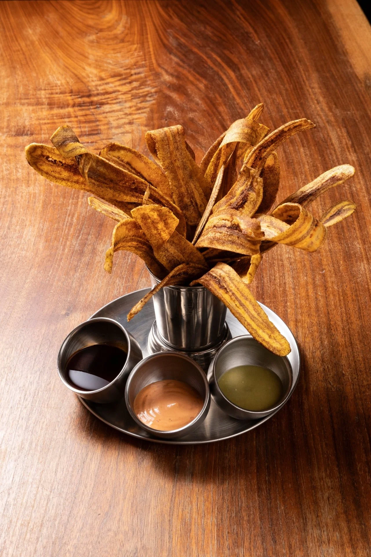Bunch of banana peels in a glass vase on a round tray with four small metal bowls containing different colored liquids on a wooden table.