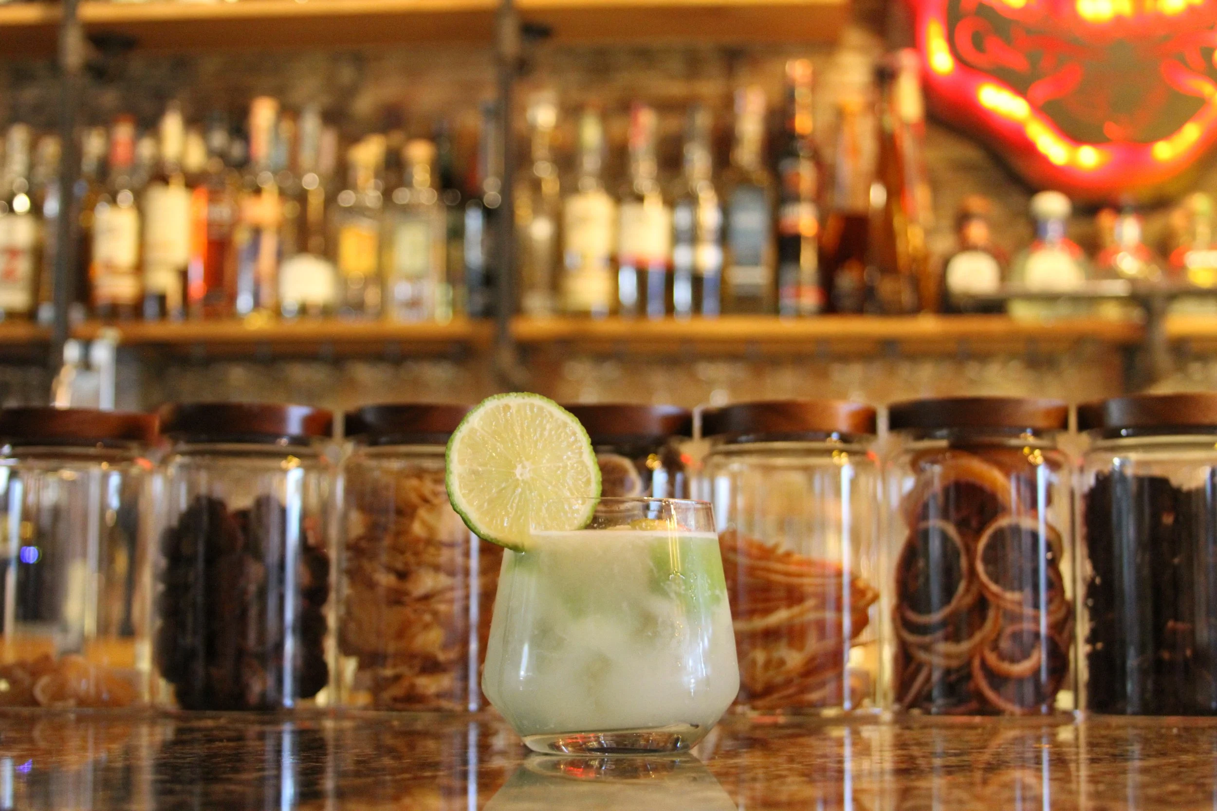 A glass of cocktail garnished with a lime slice, with a bar and liquor bottles in the background.