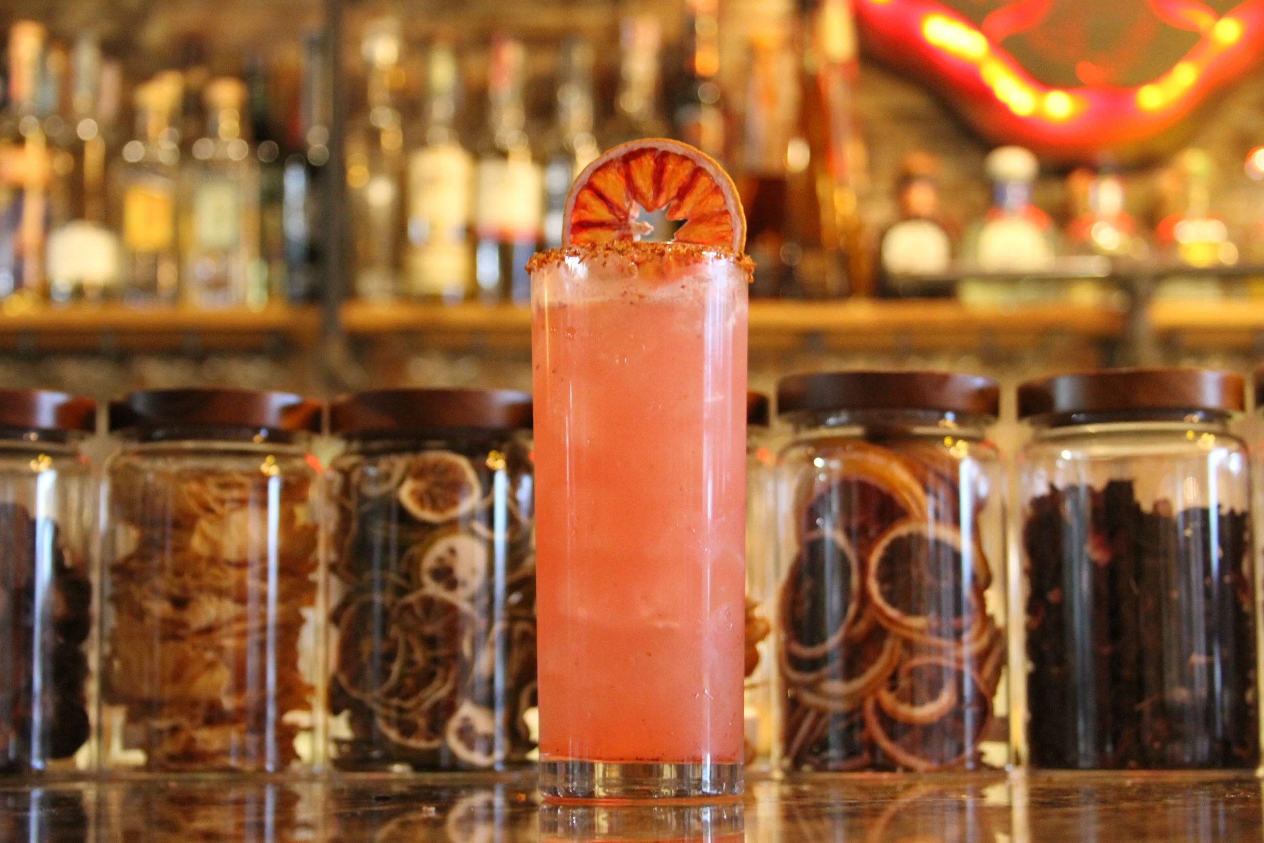 A pink cocktail with a slice of dried orange garnish on top, placed on a bar counter with blurred liquor bottles in the background.