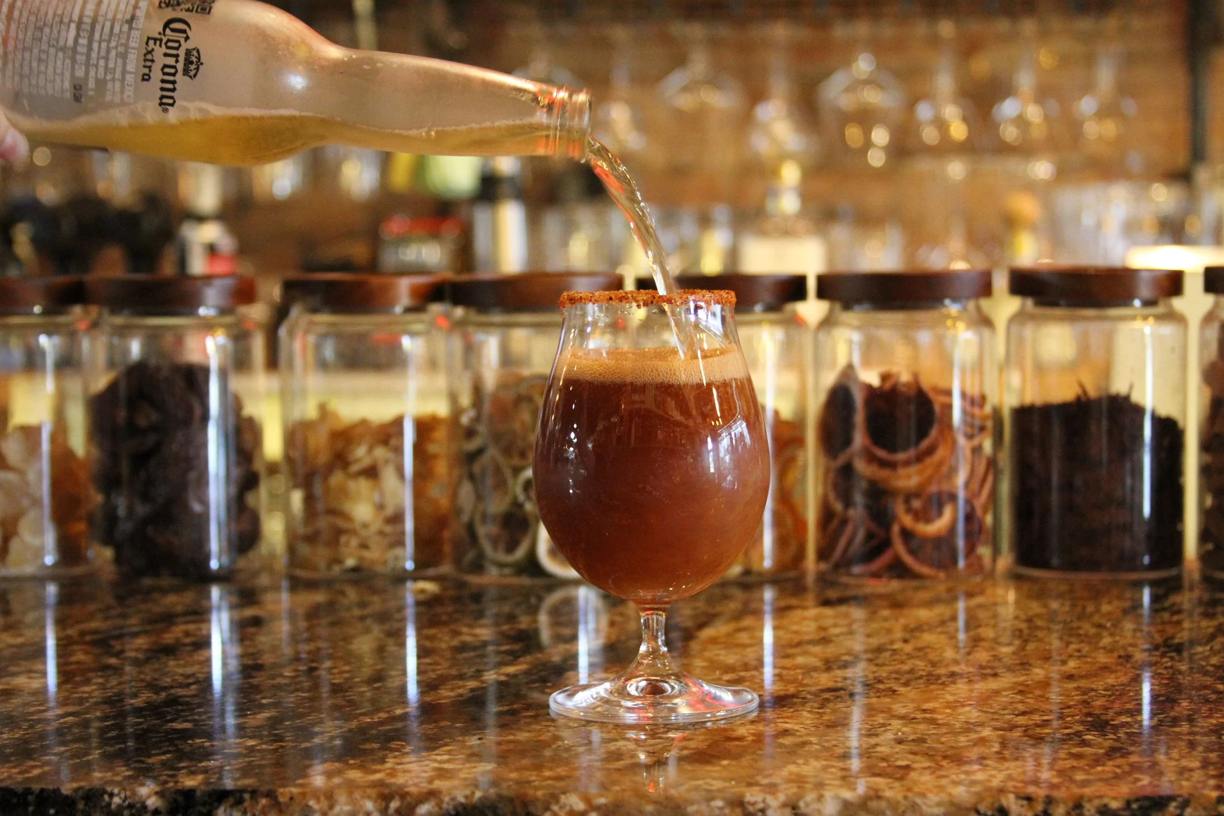 A glass of dark beer with a salted rim on a bar counter, with shelves of bottles and jars filled with liquor and spices in the background.