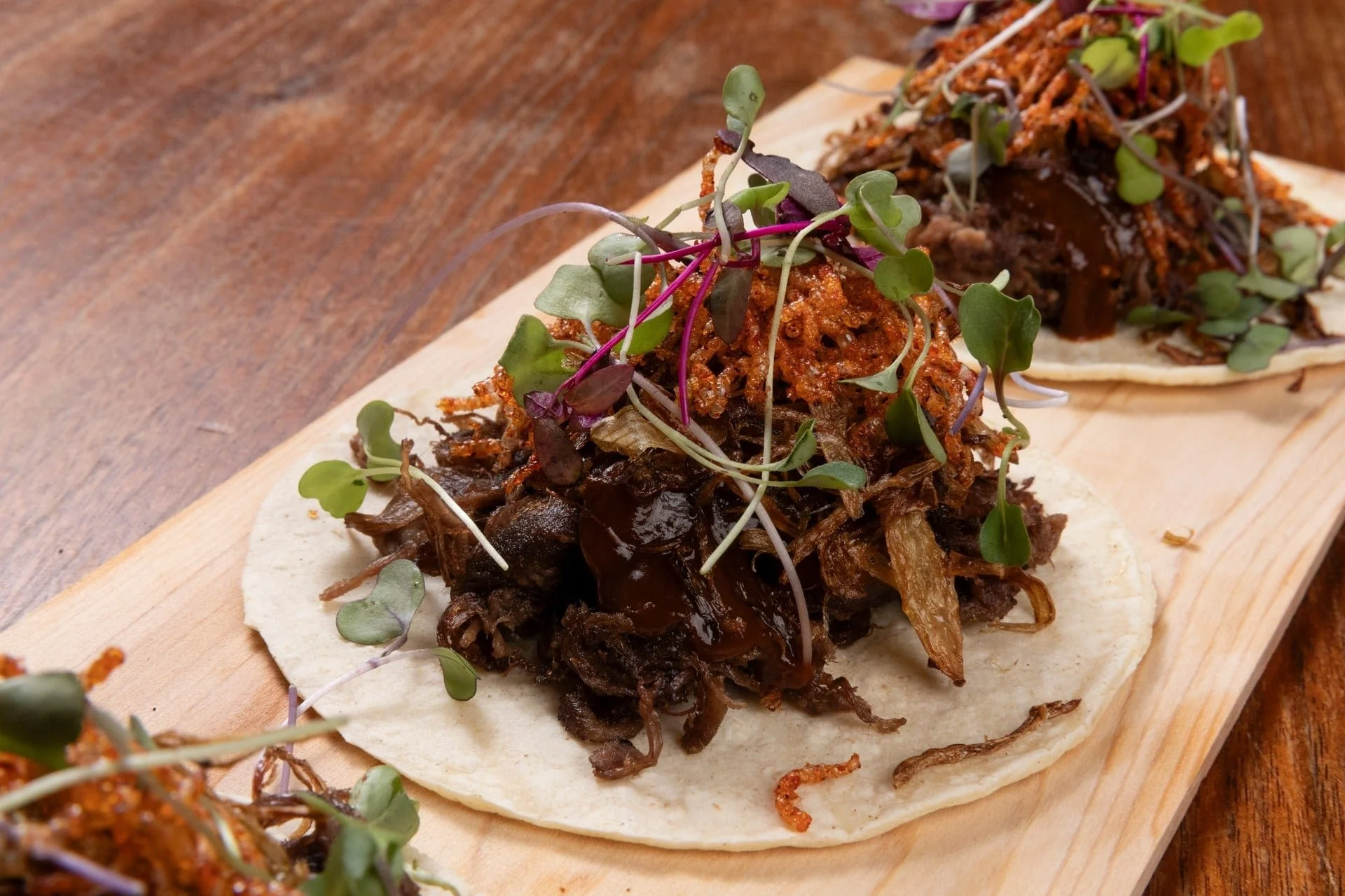 Close-up of a taco topped with shredded meat, microgreens, and spicy sauce on a wooden serving board.