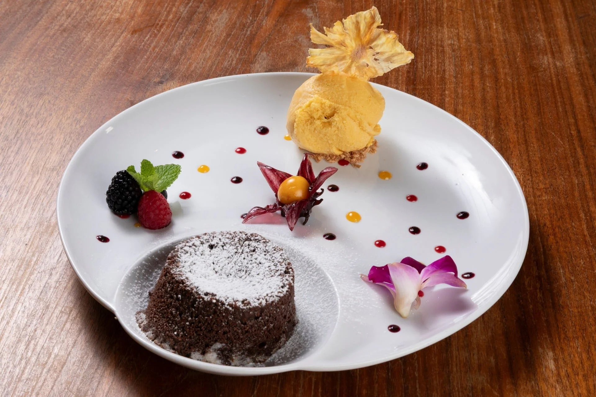 A white, leaf-shaped plate of dessert with chocolate cake, fresh berries, a scoop of mango sorbet, and a pink flower on a wooden table.