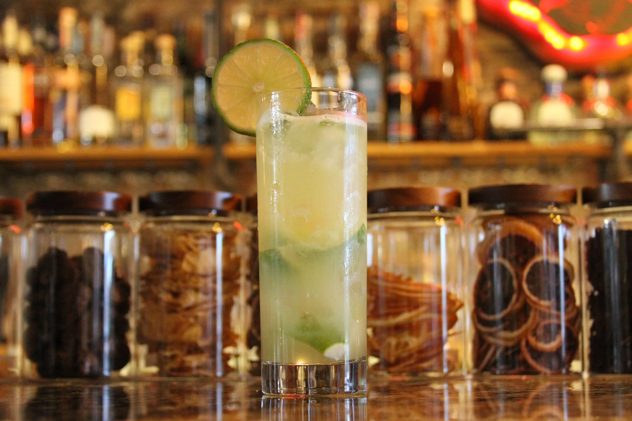 A tall glass of a lime cocktail with a lime wedge garnish on the rim, set on a bar counter with a blurred background of liquor bottles.
