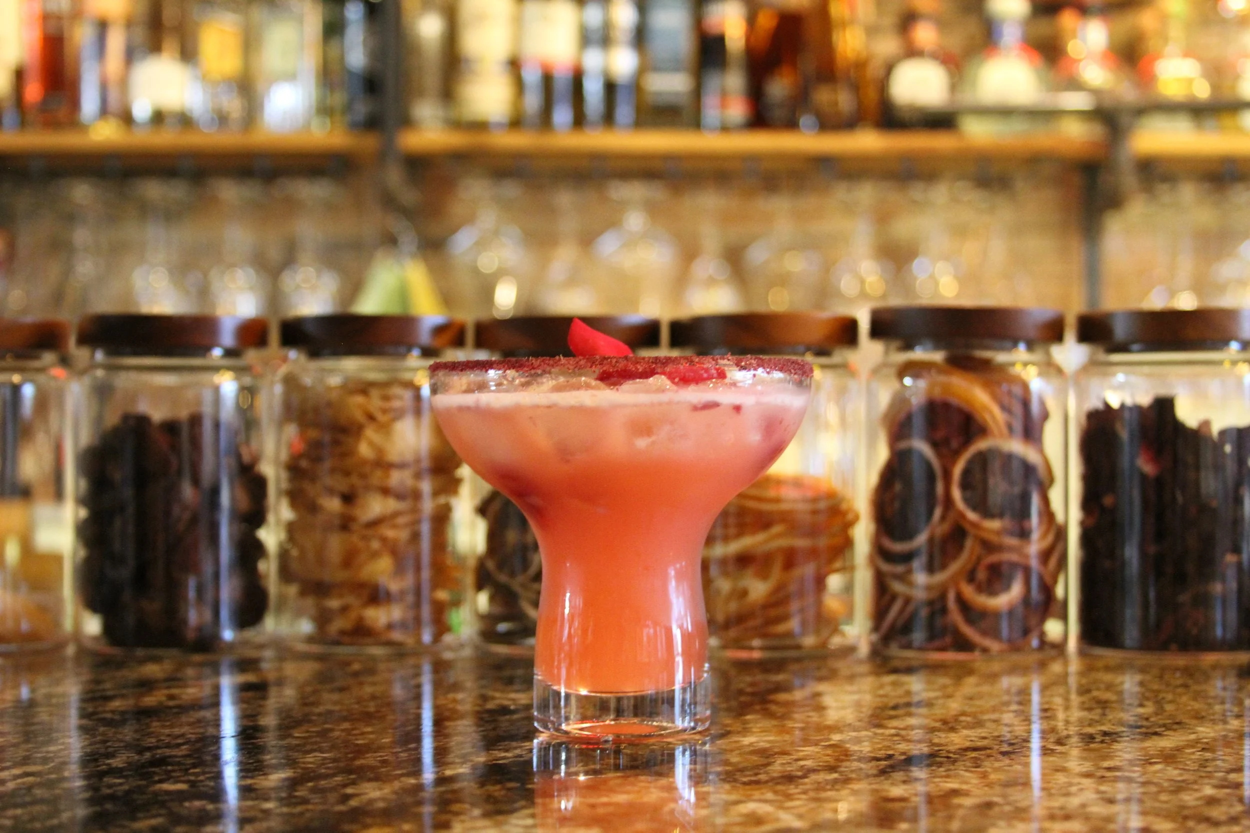 Pink cocktail in a tall glass with a red garnish, set on a bar counter with jars and liquor bottles in the background.