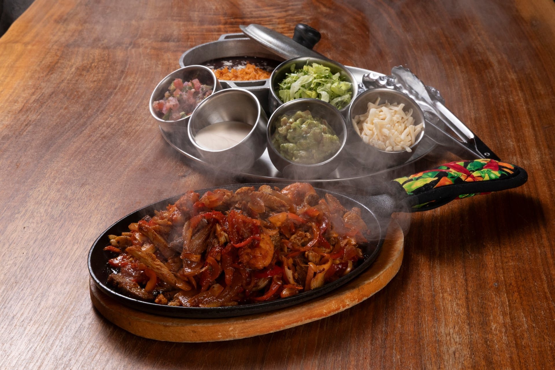 Sizzling fajita platter with cooked vegetables and grilled meat, served on a hot cast iron skillet on a wooden table, with various toppings and condiments in metal bowls in the background.