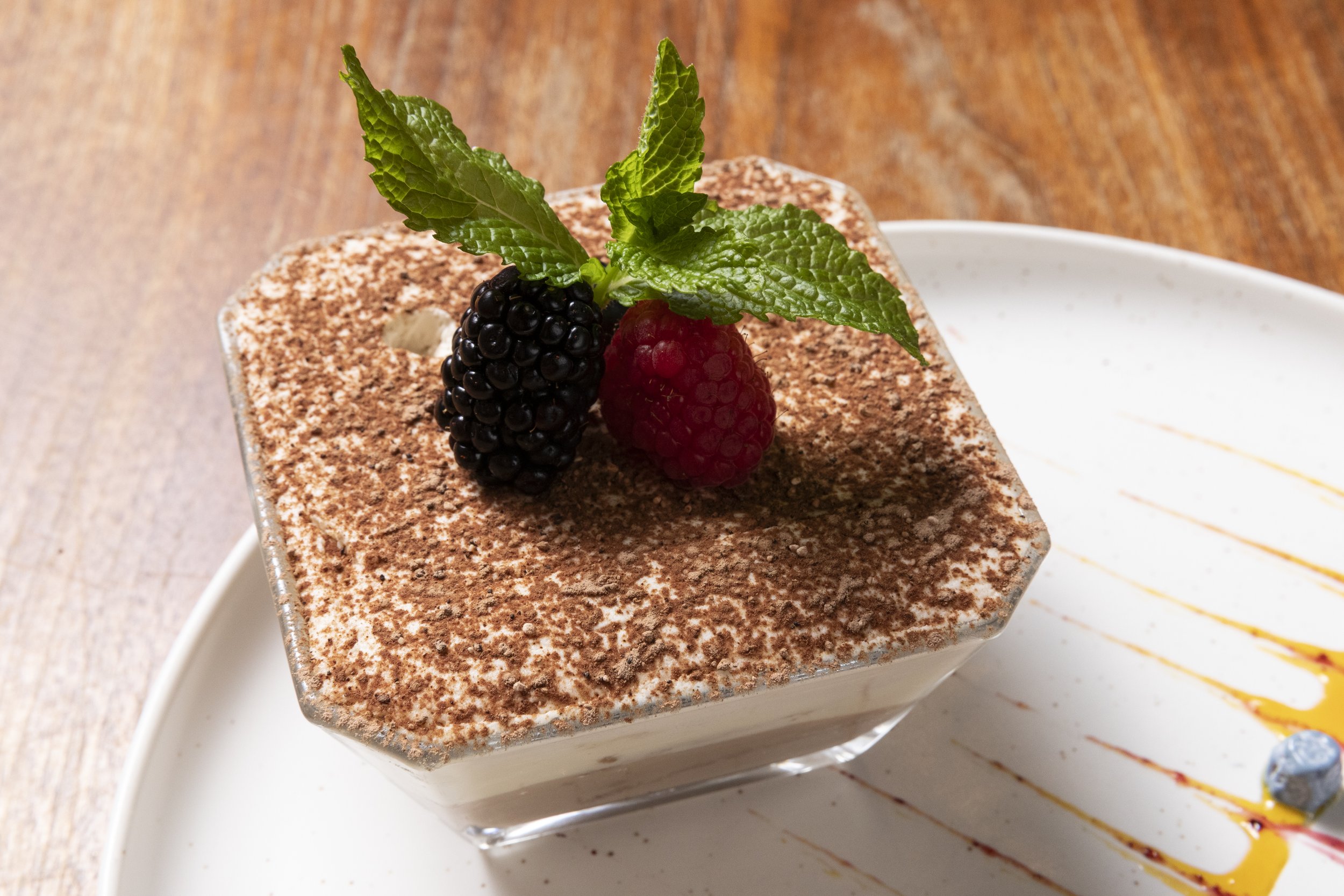 A square serving of layered dessert topped with fresh blackberries, raspberries, mint leaves, and cocoa powder, served on a white plate.