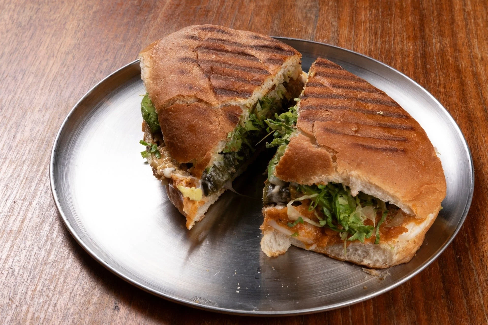 A sandwich cut in half on a round metal plate, showing lettuce, cheese, and meat inside toasted bread with grill marks, placed on a wooden table.