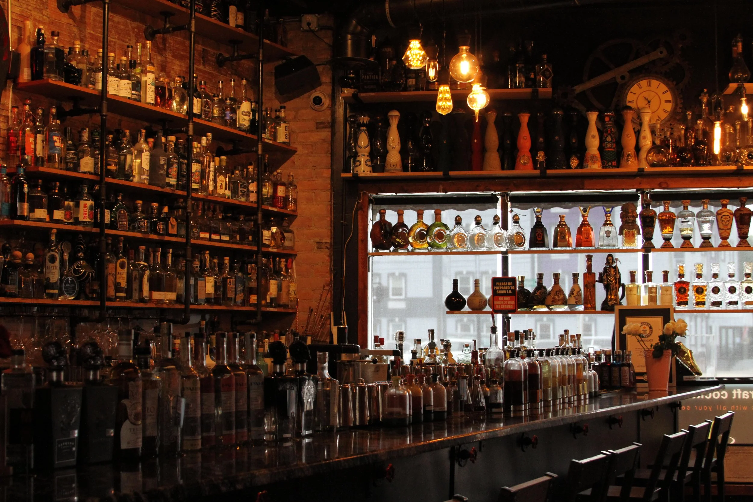 A bar with shelves filled with various liquor bottles and glasses, illuminated by warm hanging lights, with a window behind displaying a cityscape.