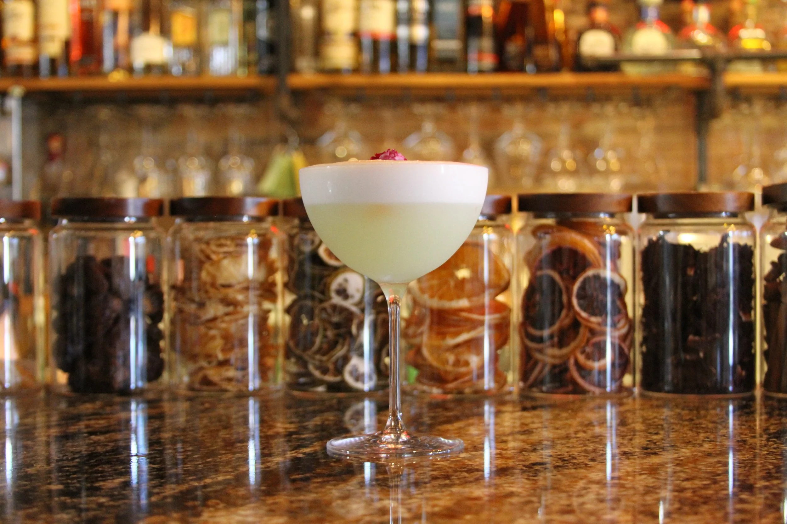A cocktail in a coupe glass with a pink garnish, set on a bar counter with jars of dried herbs or spices and shelves of bottles in the background.