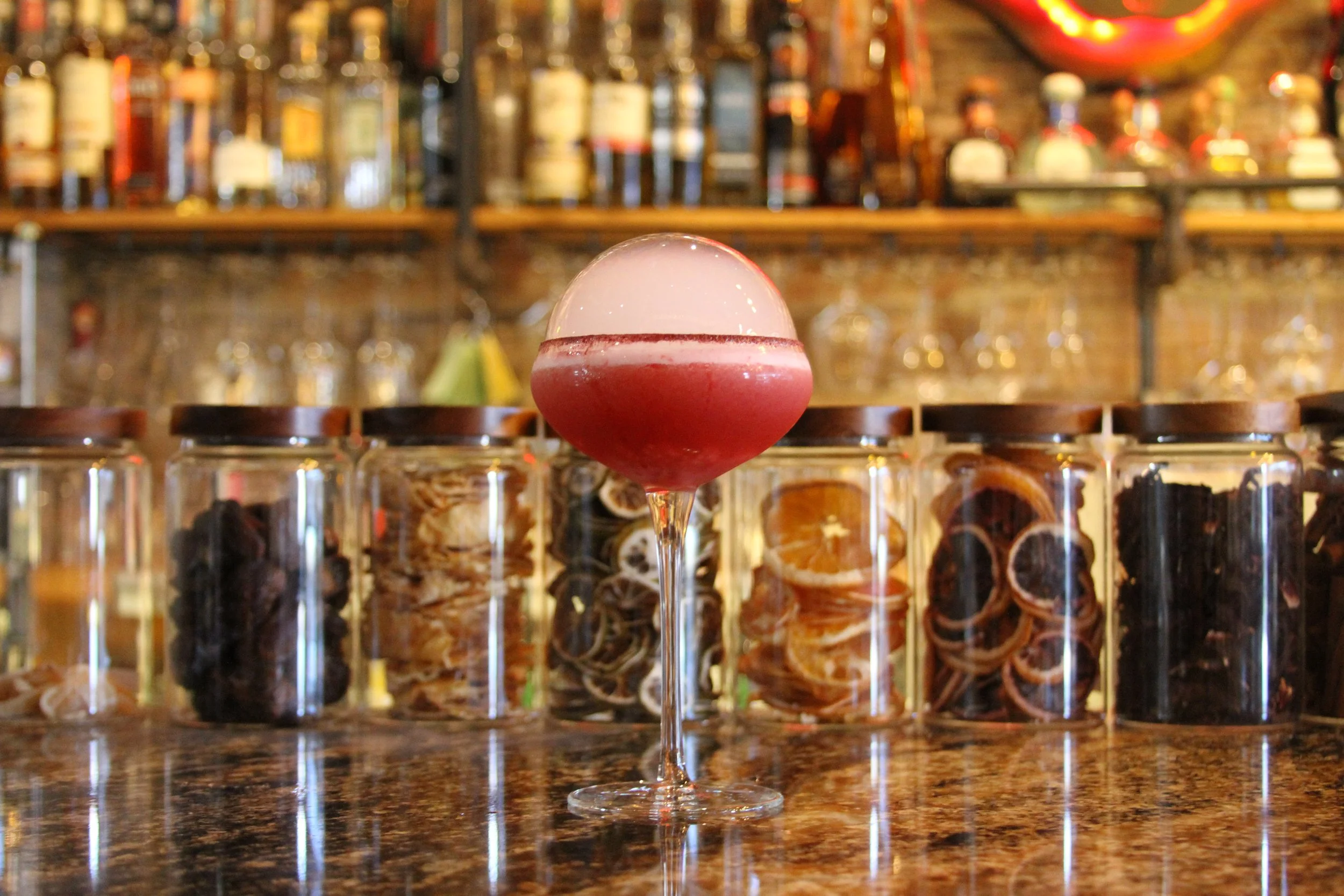 A cocktail in a large wine glass with a frothy top, placed on a bar counter. The background features a blurred bar with various bottles and glasses, creating a vibrant and cozy atmosphere.