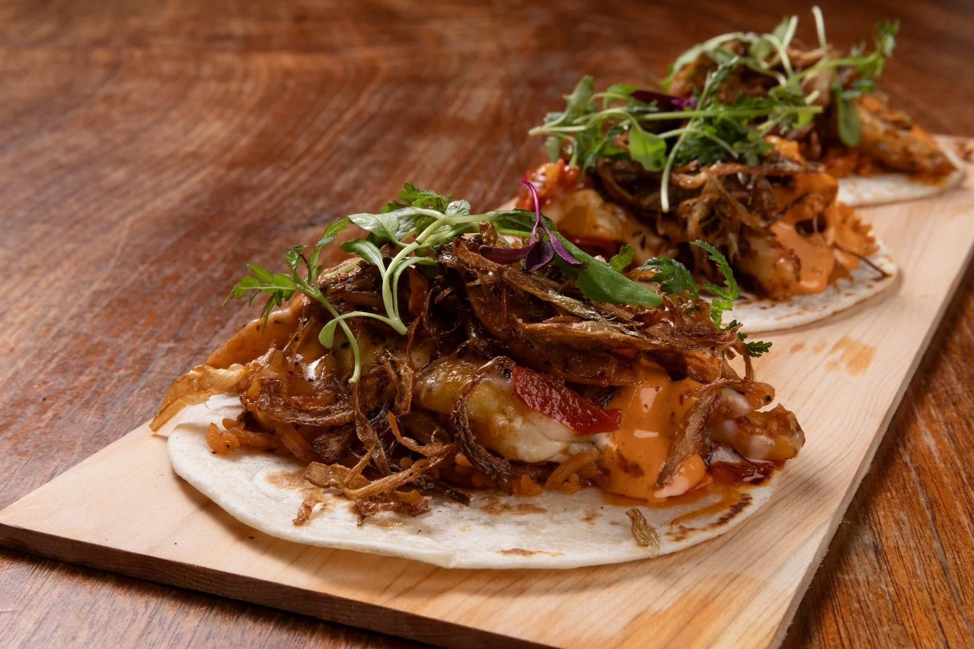 Two tacos on a wooden board filled with shredded meat, fresh greens, and sauce.