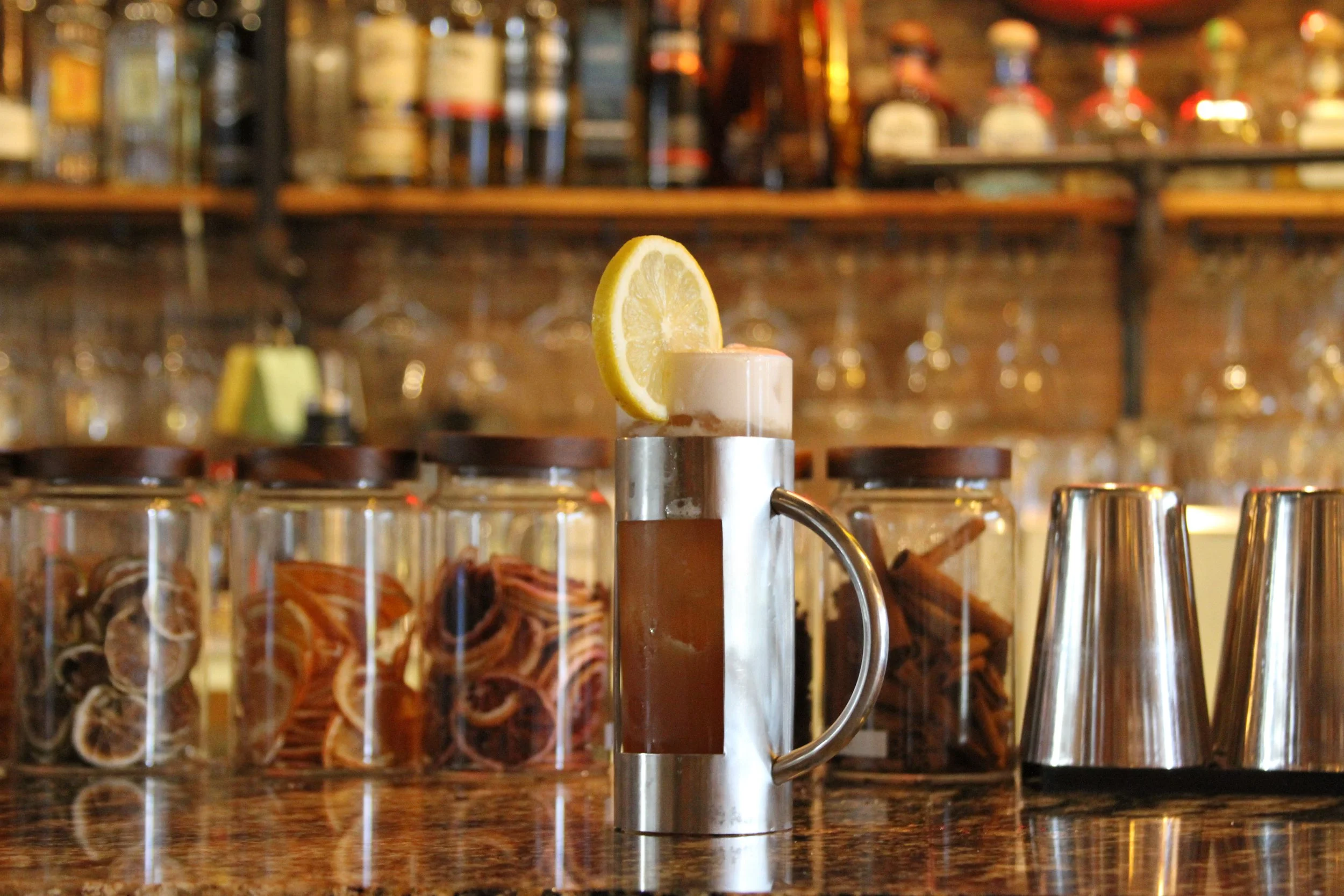 A metal lemon squeezer with a lemon wedge on top, placed in front of a row of glass jars filled with ingredients, with a bar or kitchen counter behind.