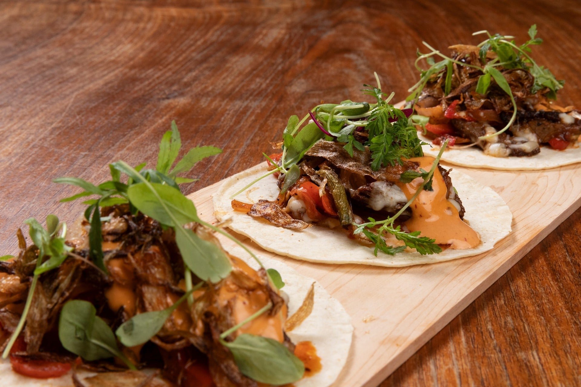 Close-up of three tacos on a wooden serving board with various toppings including greens and sauce, on a wooden table.
