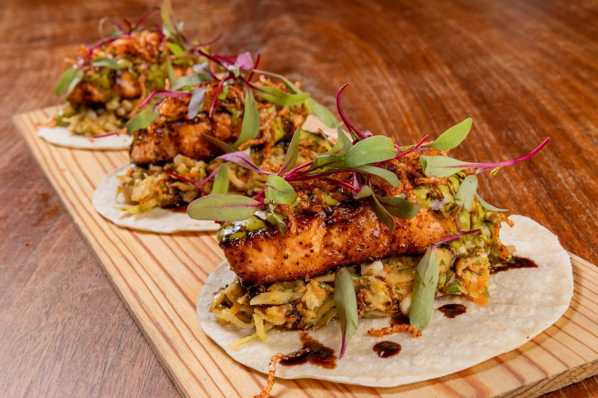 Close-up of three fish tacos topped with microgreens on a wooden serving board, placed on a wooden table.