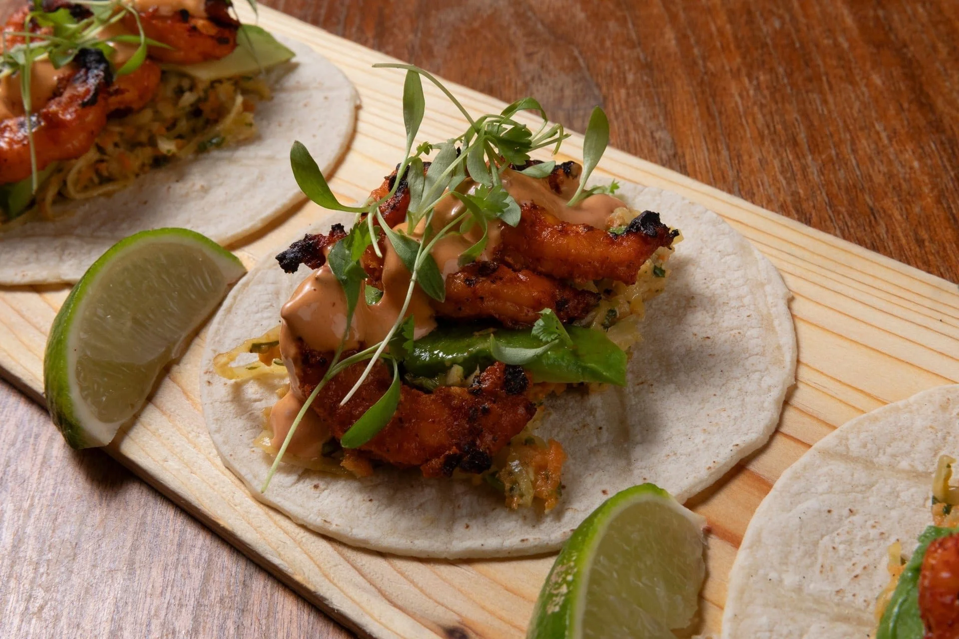 Close-up of fish tacos topped with microgreens, served with lime wedges on a wooden platter.