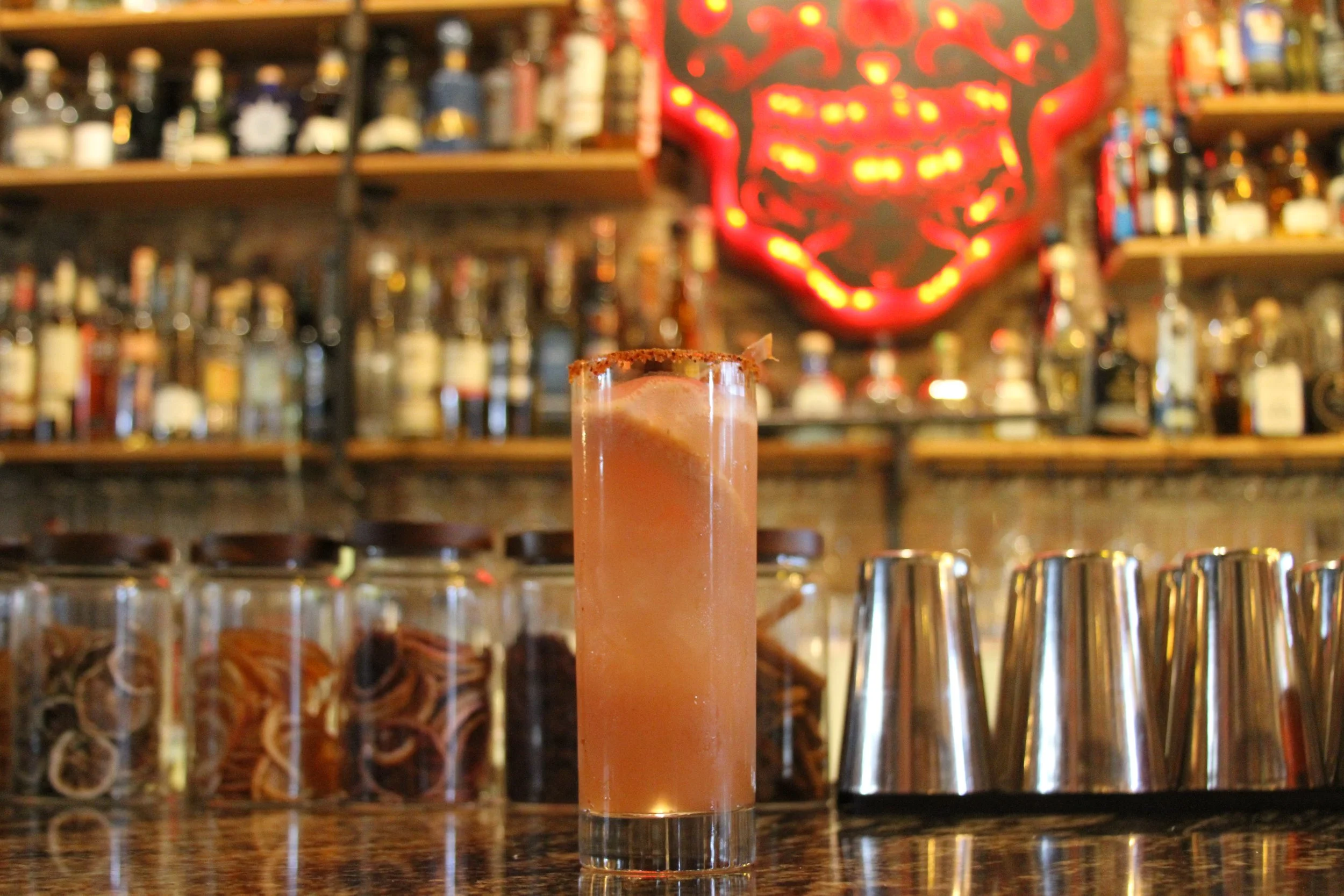 A pink cocktail in a tall glass with a salted rim, placed on a bar counter. There are blurred bottles of alcohol on shelves in the background, and a red neon face sign is visible.