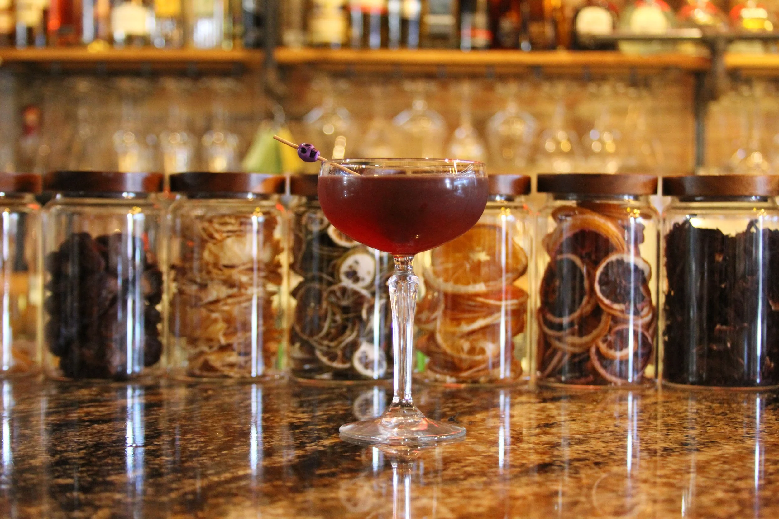A cocktail glass with a dark red drink and a small skewer with purple and black objects on top, in front of a row of glass jars filled with dried fruit slices, set on a countertop with a bar in the background.