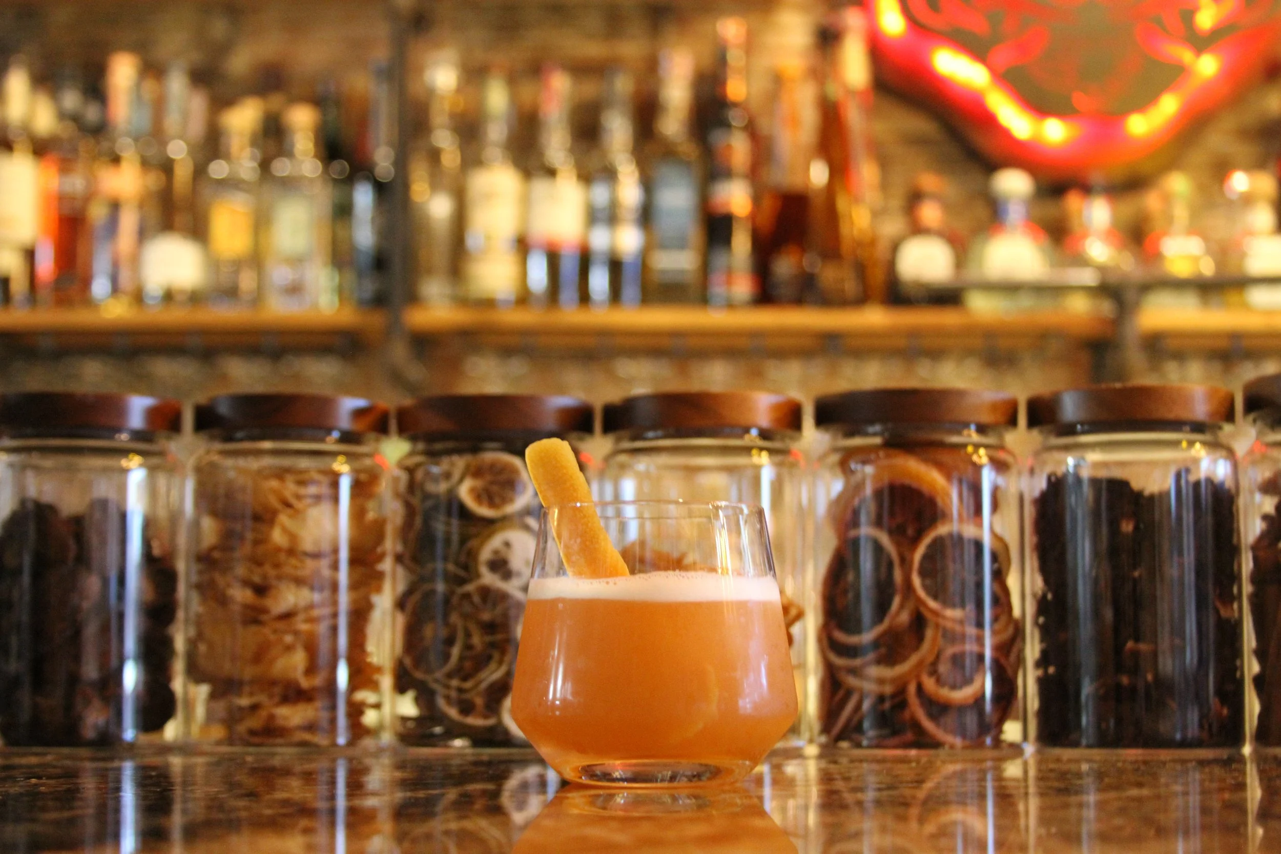 A glass of orange-colored cocktail with a straw, in front of a blurred background of shelves filled with bottles and a neon sign, in a bar setting.