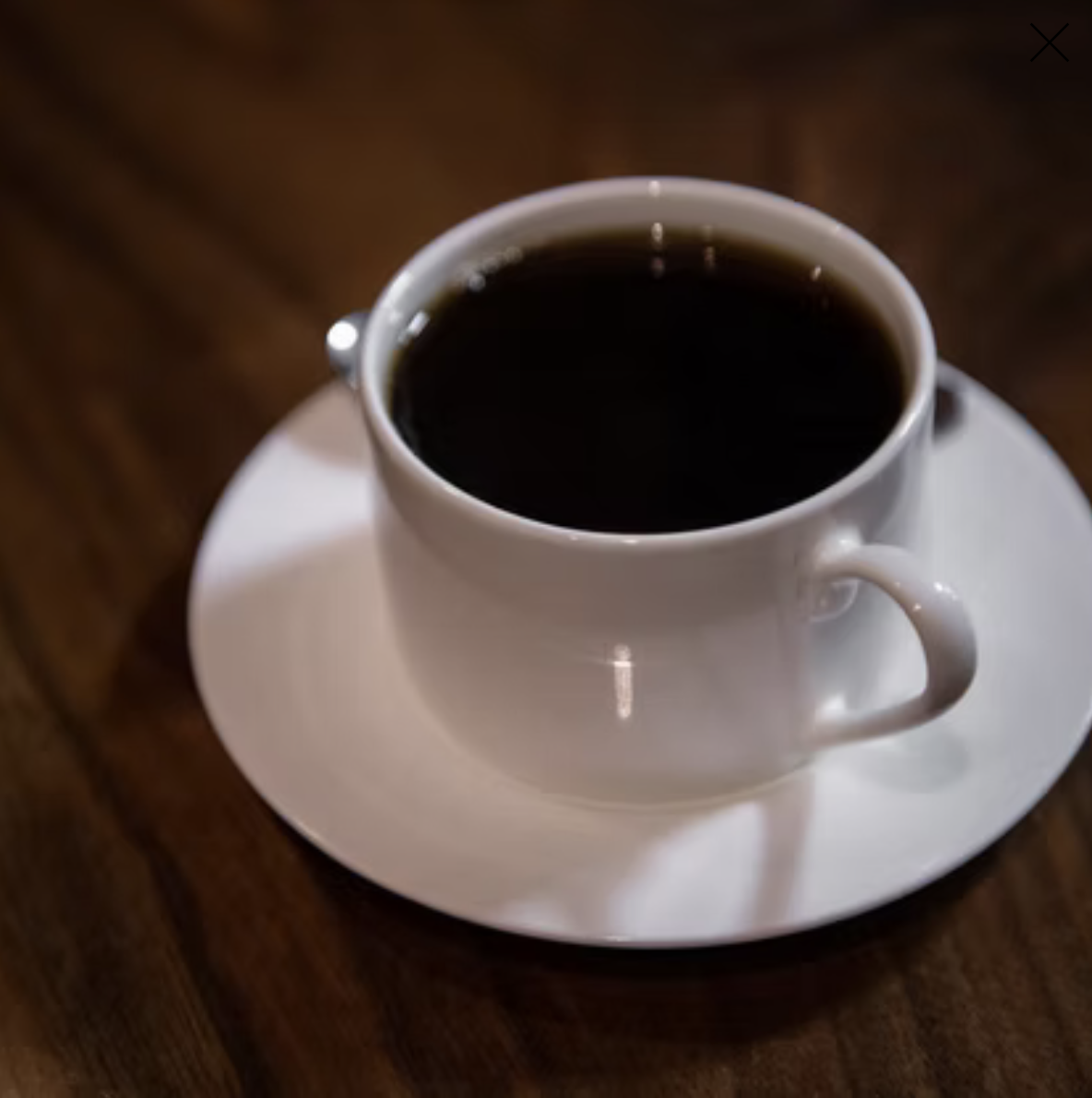 A white coffee cup filled with black coffee on a matching white saucer, placed on a dark wooden table.