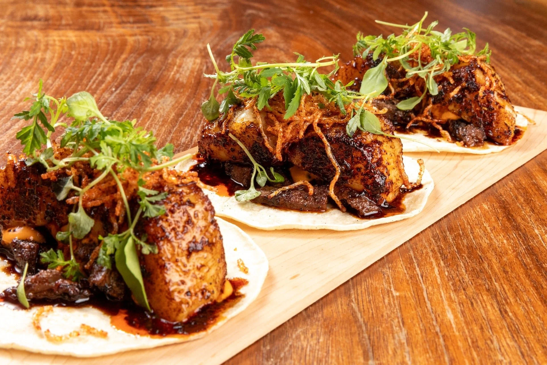 Three tacos filled with meat, topped with herbs and sauce, served on a wooden board.