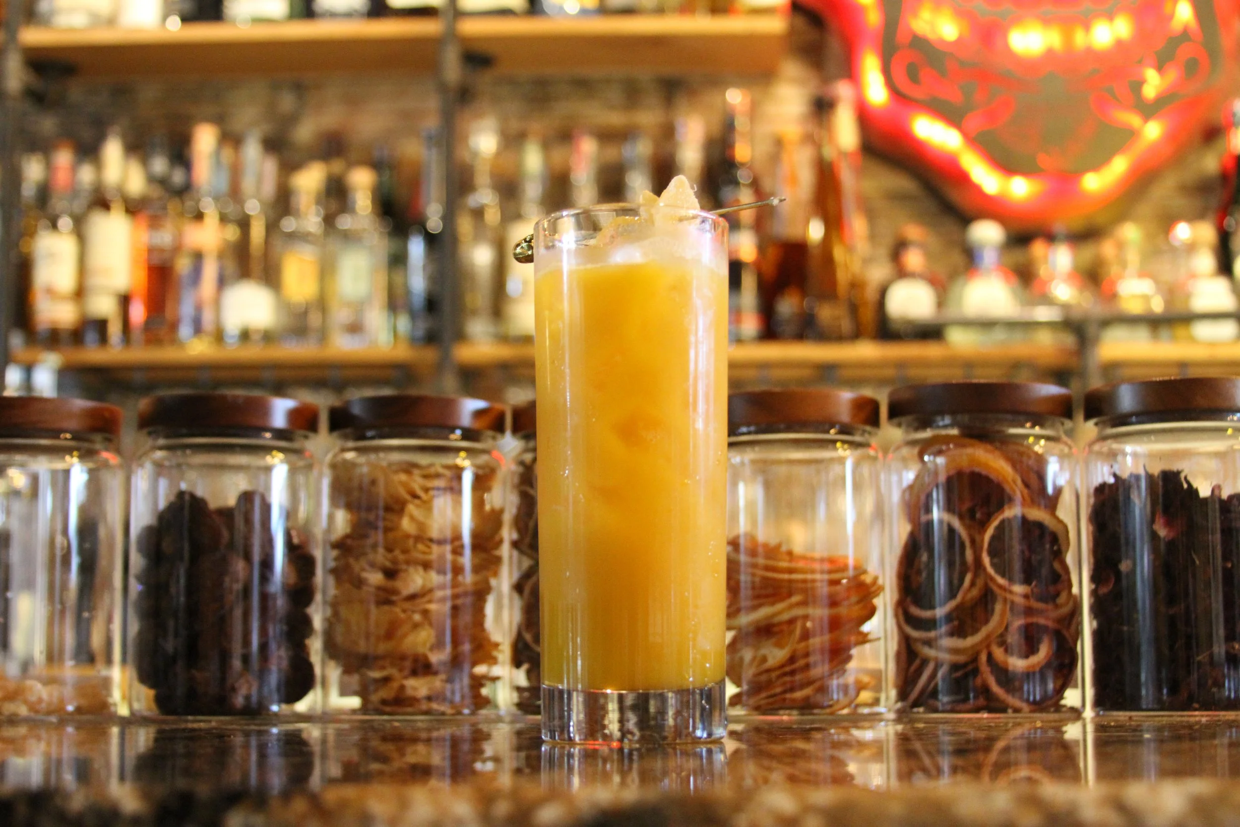 A tall glass of yellow cocktail with ice and a cherry on a skewer, placed in front of a row of glass jars filled with dried fruit, in a bar setting with bottles on shelves in the background.