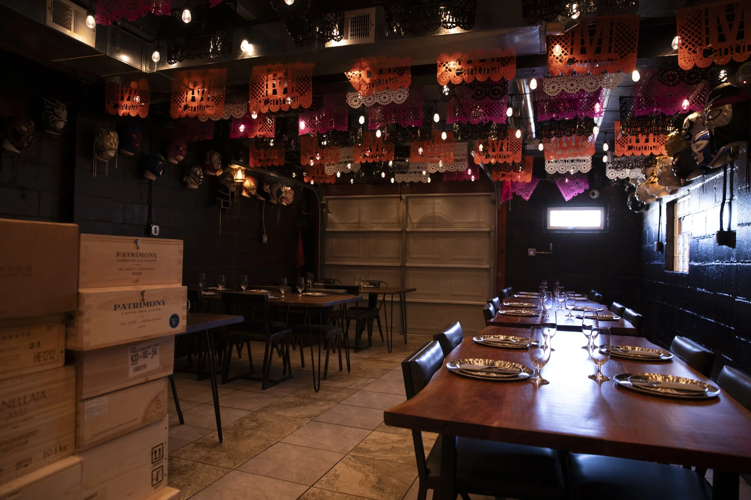 A dimly lit restaurant decorated for Cinco de Mayo with pink, black, and white paper banners hanging from the ceiling. There are masks displayed along the wall, a wooden table set for dining with wine glasses and plates, and a window on the back wall letting in natural light.