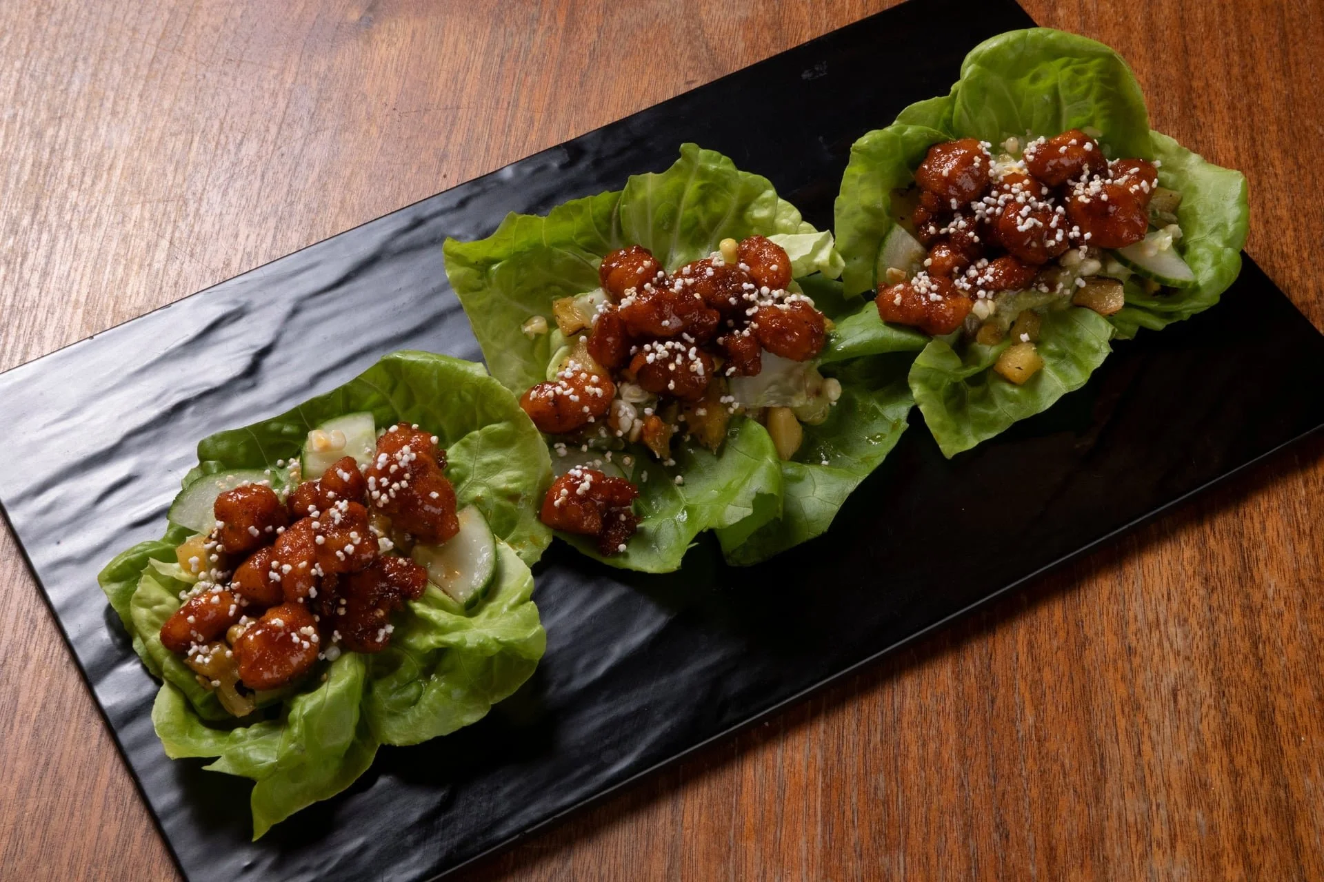 Four lettuce wraps filled with chicken and topped with white sesame seeds on a black rectangular plate, on a wooden table.