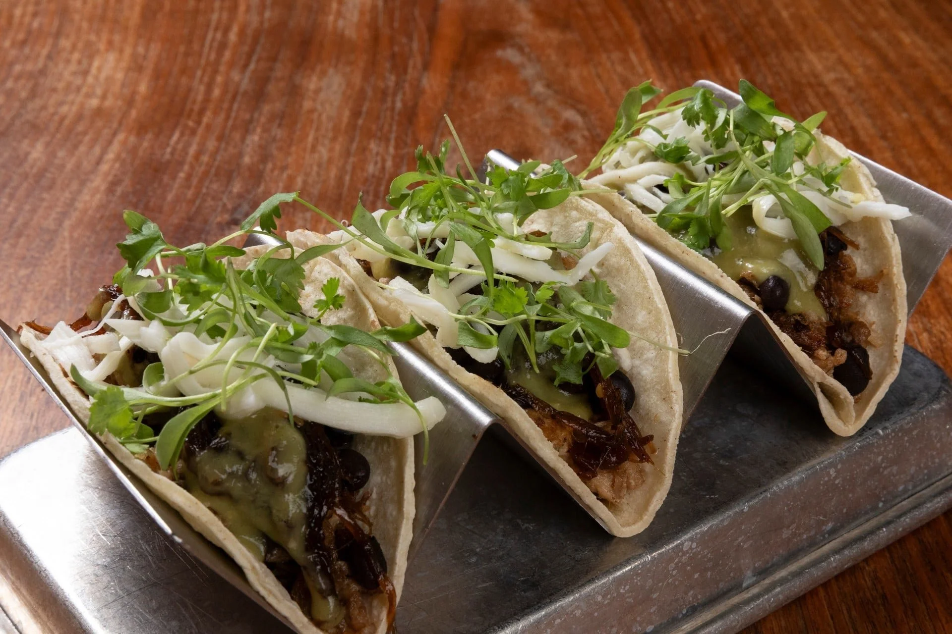 Three tacos with greens, shredded cheese, black beans, and melted cheese in a metal taco holder on a wooden table.