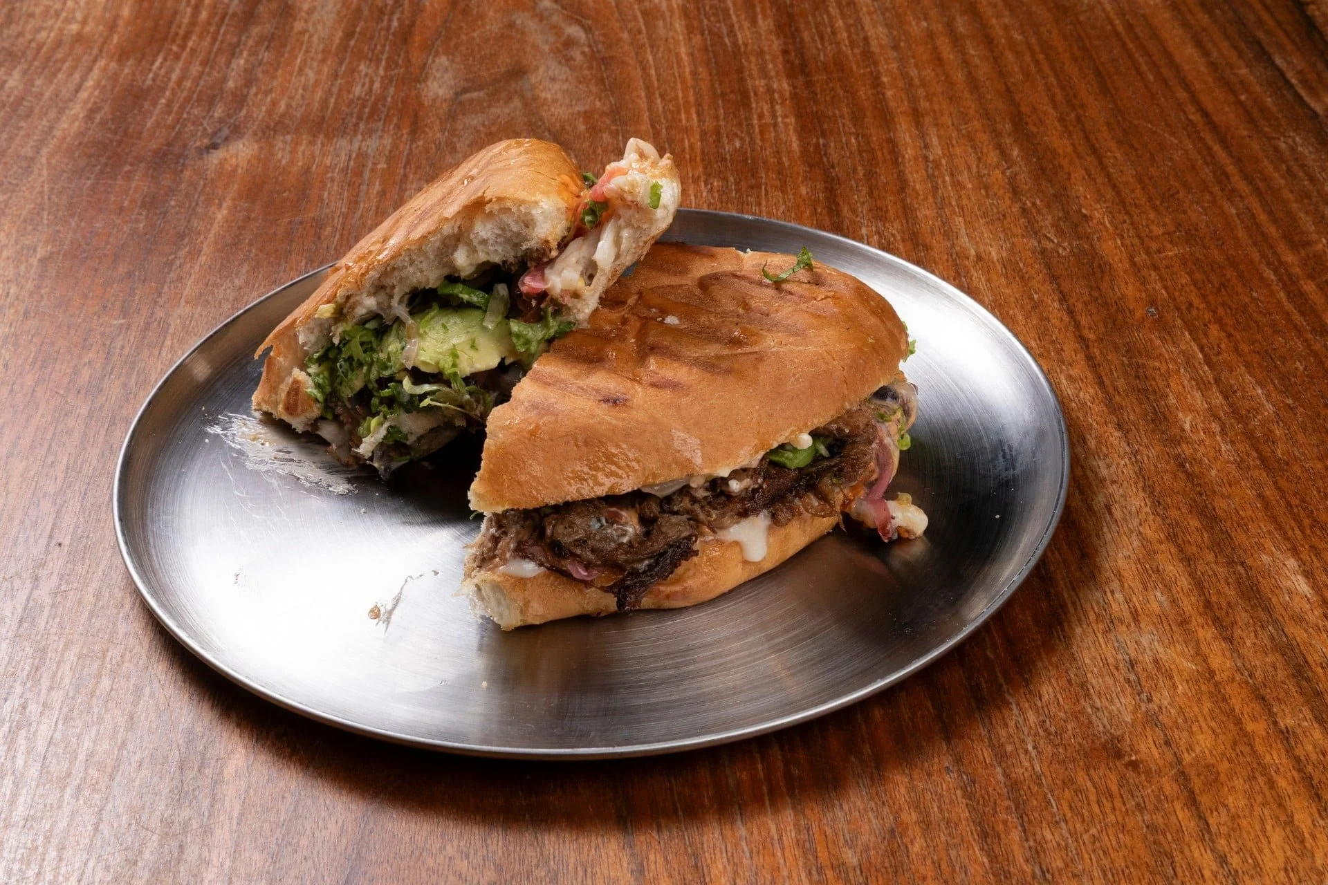 A silver platter holding a large sandwich with slow-cooked beef, pickles, onions, lettuce, and sauce, on a wooden table.