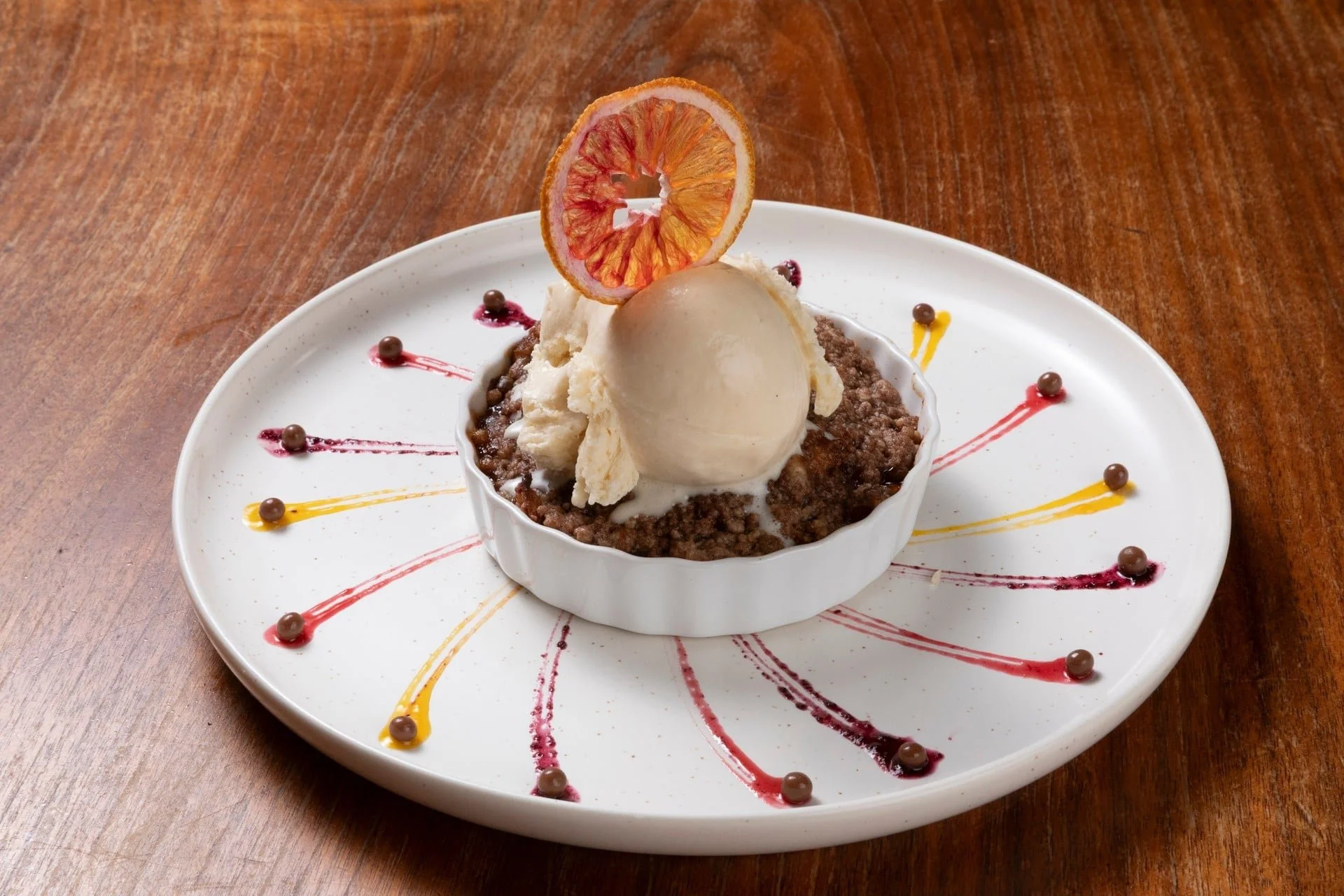Dessert with a scoop of vanilla ice cream on a chocolate crumble base, garnished with a dried citrus slice, served on a white plate with colorful sauce and chocolate pearls.