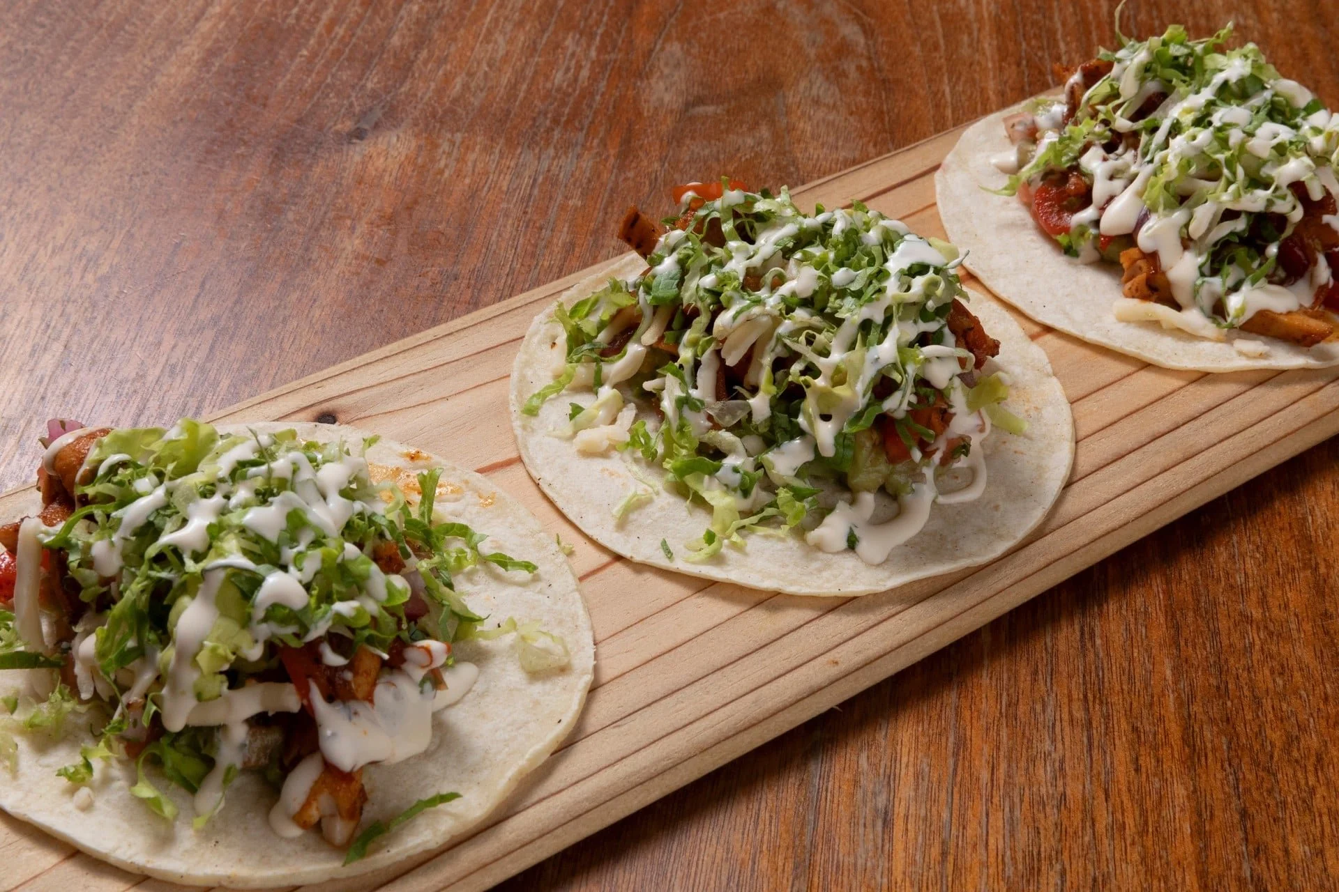 Three tacos on a wooden board topped with chopped lettuce, shredded cheese, and drizzled with white sauce.