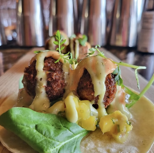 Two meatballs topped with melted cheese and garnished with fresh herbs on a bun with lettuce, served with a side of diced yellow peppers.
