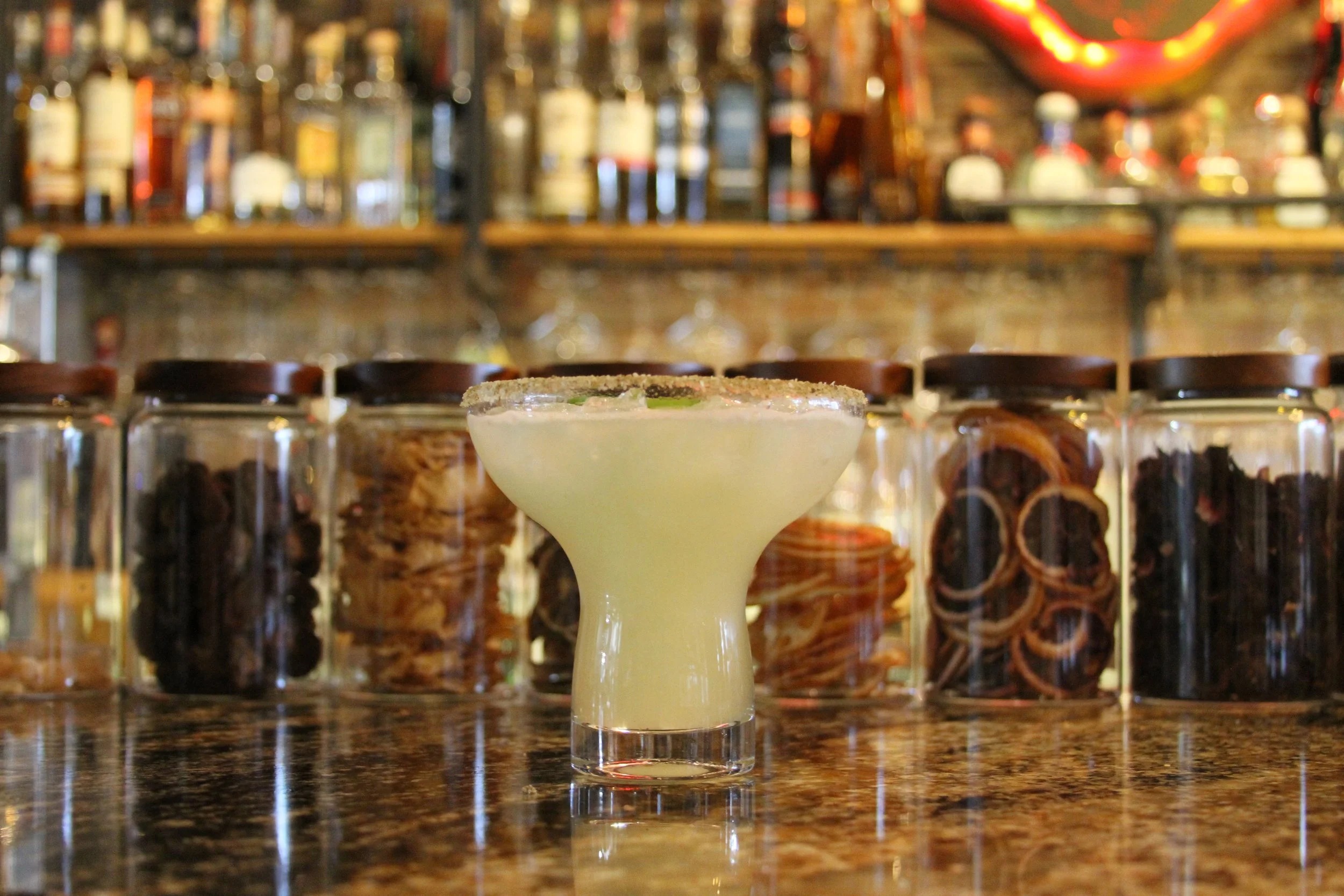 A cocktail drink in a stemmed glass with a salted rim and lime garnish, with rows of spice jars behind on a shelf in a bar setting.