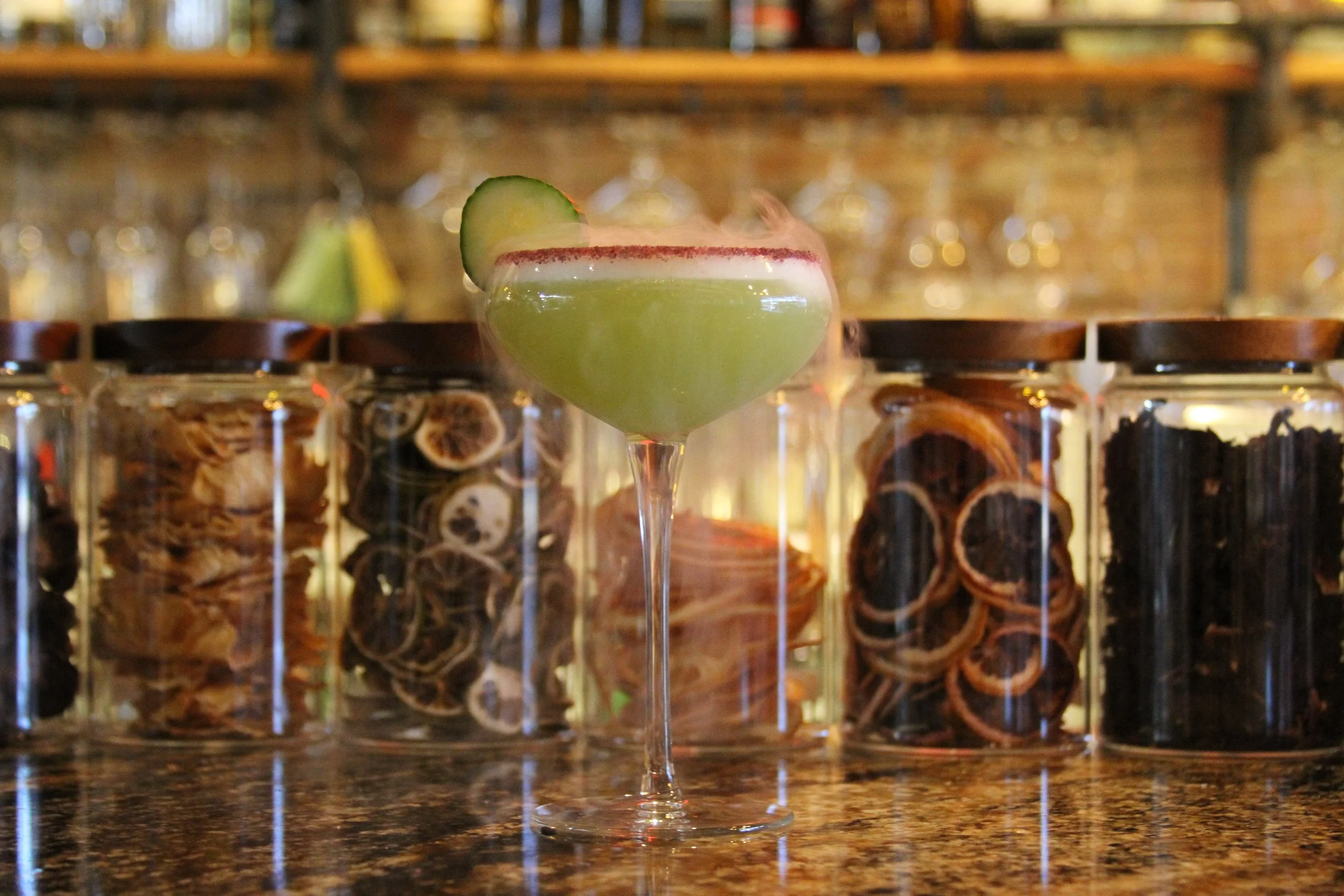 A green cocktail with a salted rim and cucumber garnish in a coupe glass, set against a bar background with shelves of jars and bottles.