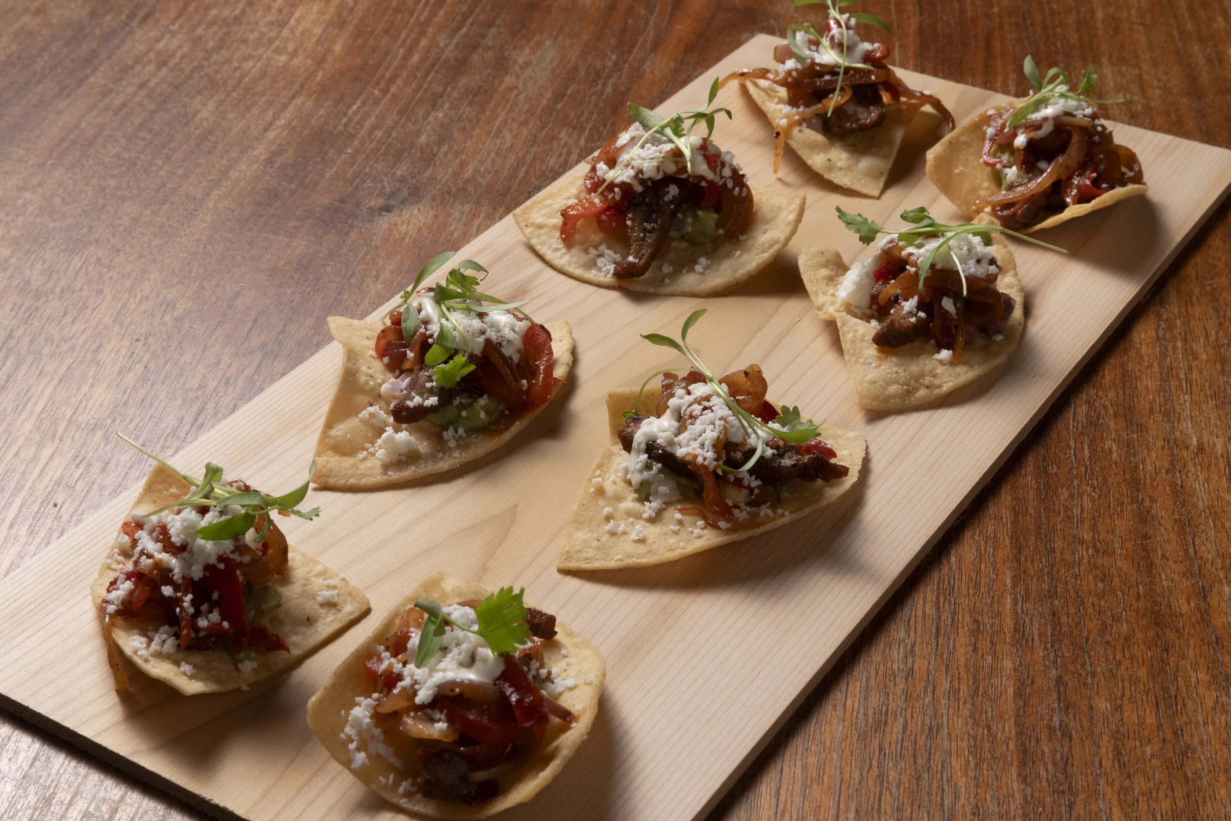 Multiple small tacos with beef, diced tomatoes, cheese, and greens on a wooden board.