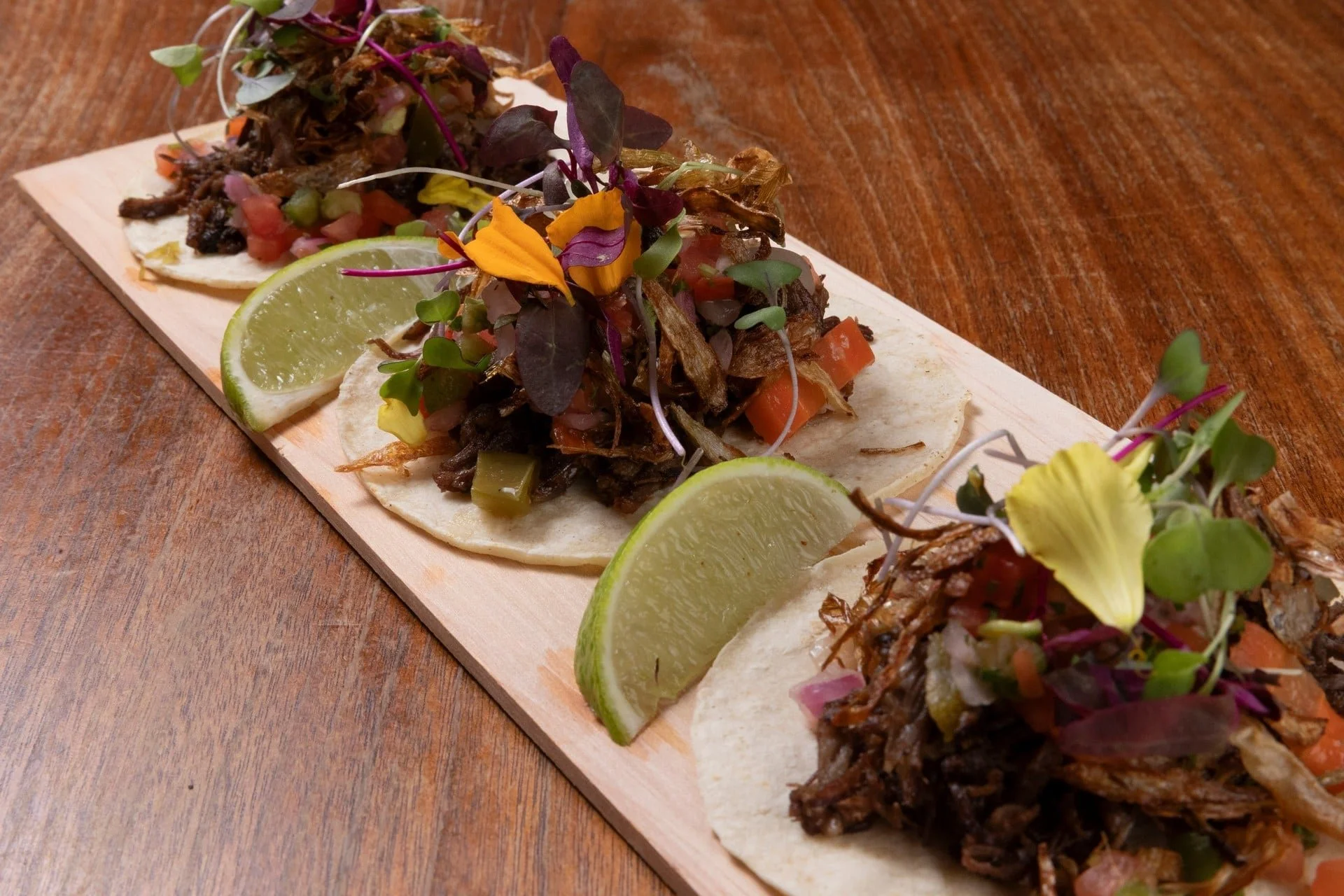 Close-up of a wooden serving board with several small tacos filled with shredded meat, chopped vegetables, and topped with microgreens and edible flower petals, garnished with lime wedges.