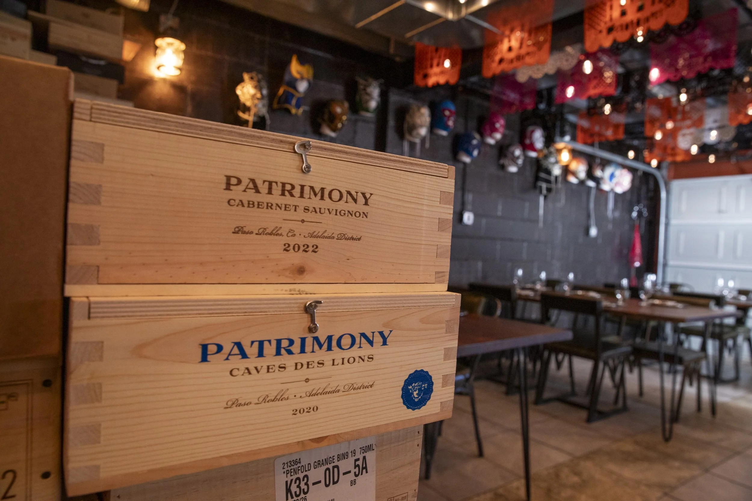 A wooden box with the words 'PATRIMONY CABERNET SAUVIGNON 2022' printed on it is in the foreground. The background shows a restaurant or bar interior with tables, chairs, and hanging decorations, including colorful masks and paper banners.
