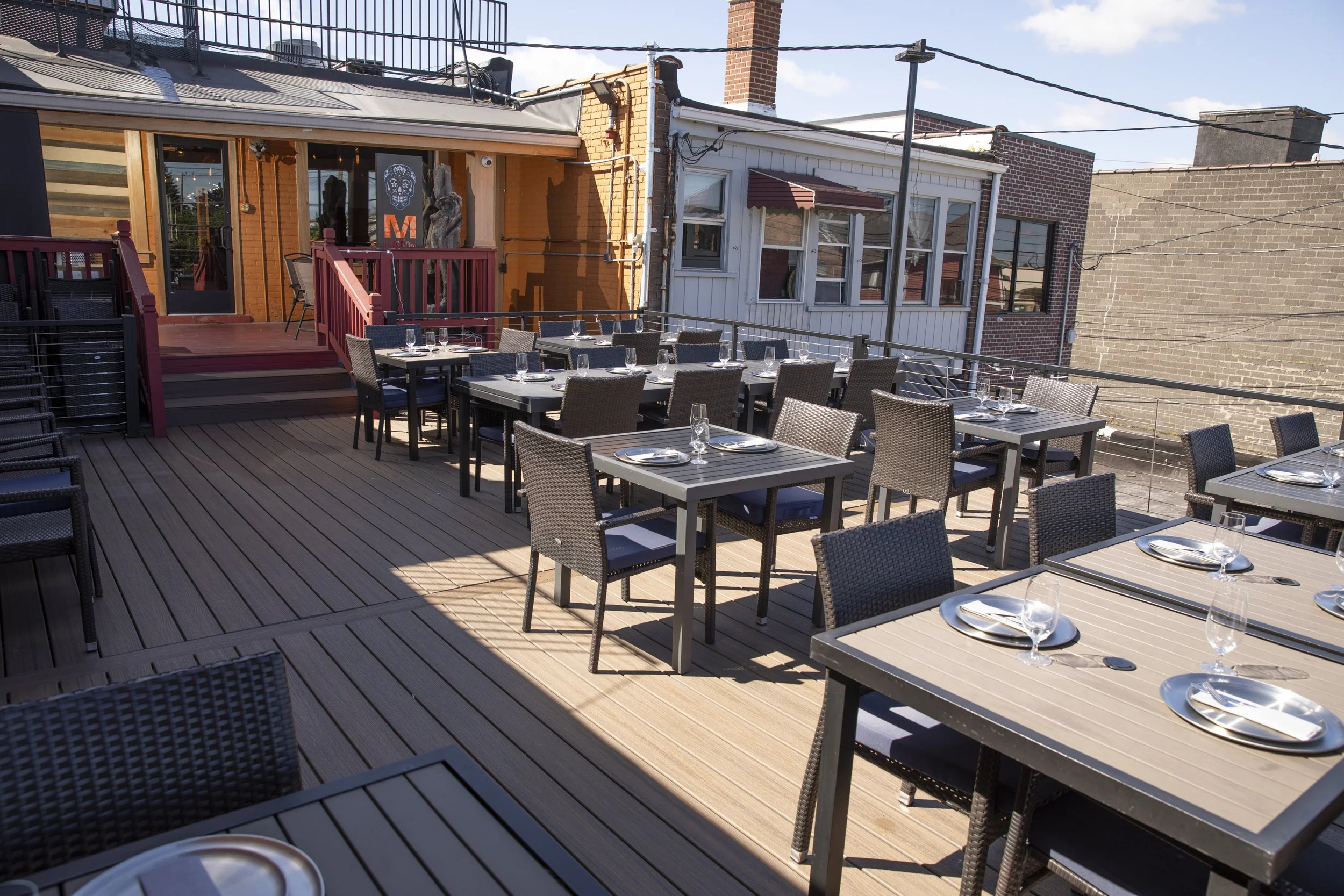 An outdoor rooftop dining area with multiple tables set with glasses and plates, surrounded by chairs, against a backdrop of brick and wooden buildings under a clear blue sky.